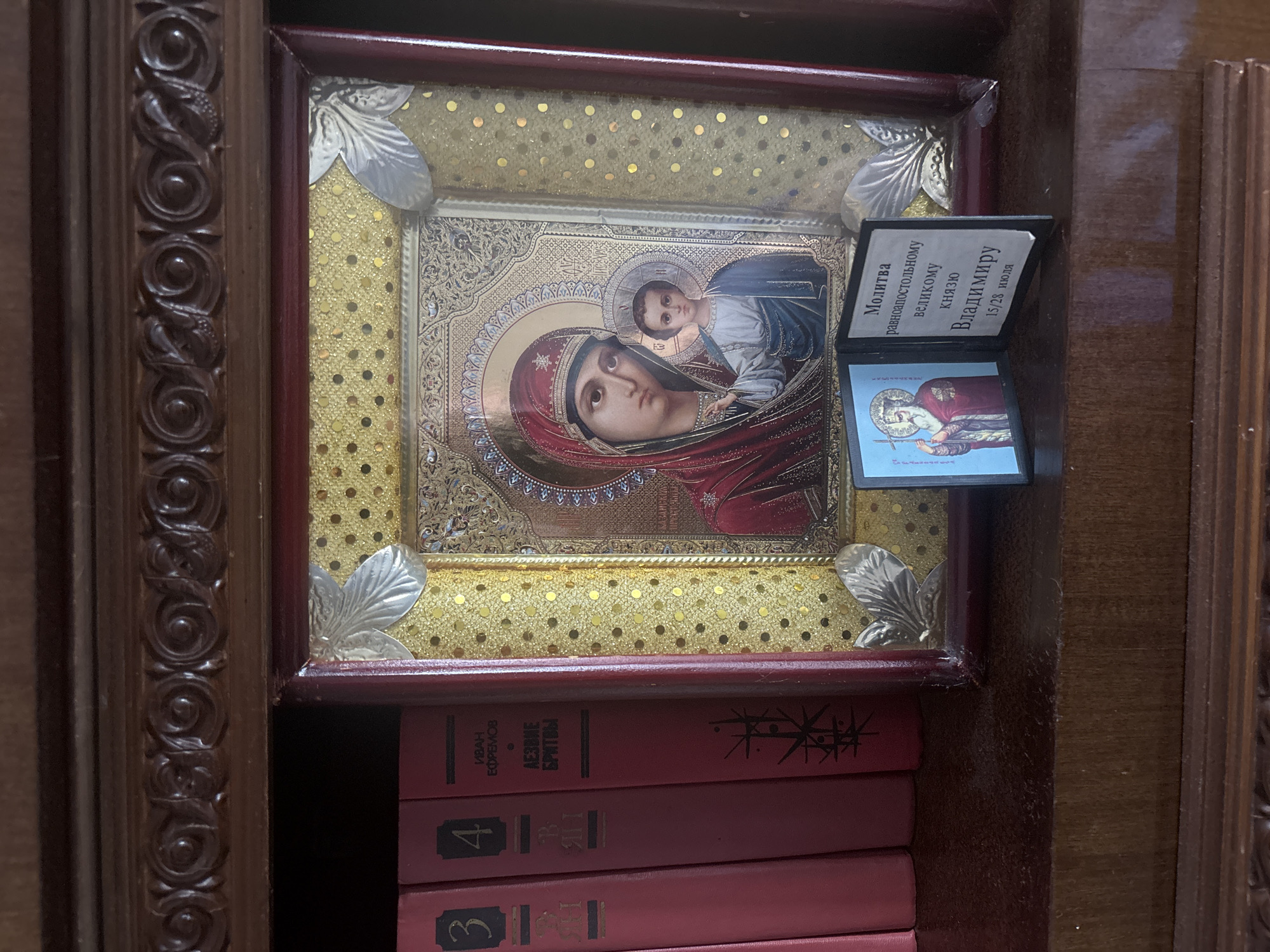 A religious icon of Mary and the Christ child is displayed on a bookshelf in an interior setting. The icon is framed with red and gold, adorned with ornate patterns and silver leaf accents. Beside the icon, a small, open booklet displays an image of a bearded man in religious attire and text in a foreign language. Three red-bound books are visible to the left of the icon, with numbers and text printed on their spines. The bookshelf appears to be made of dark wood with decorative carvings along the top edge. The overall scene suggests a personal collection or a place of worship. The location is identified as Minsk, Belarus.