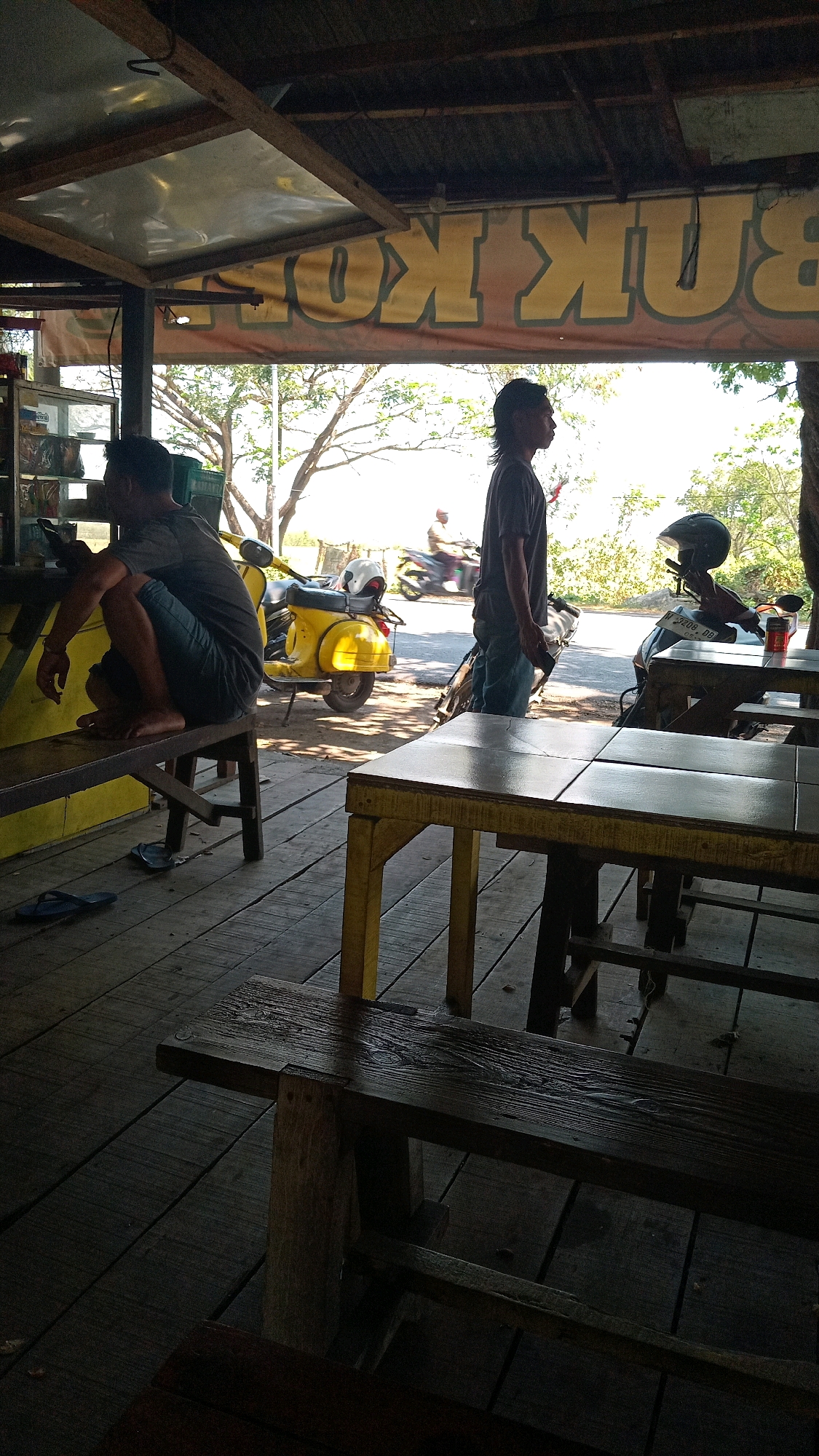 A man in a dark t-shirt and jeans stands facing right, his profile visible, in front of a shop. To his left, another man, wearing a dark t-shirt and shorts, squats on a wooden bench, looking at his phone. A yellow scooter is parked behind the squatting man, with a white helmet on its seat. A black scooter with a helmet on its seat is parked further right, behind a wooden table. The scene appears to be an outdoor establishment, possibly a roadside stall, in Ambeng Ambeng Watangrejo, Indonesia. The ground is made of wooden planks, and there are wooden tables and benches. A banner with large yellow letters is visible in the background, with the word "BUNKOT" partially legible. The overall lighting suggests it is daytime, with sunlight casting shadows.