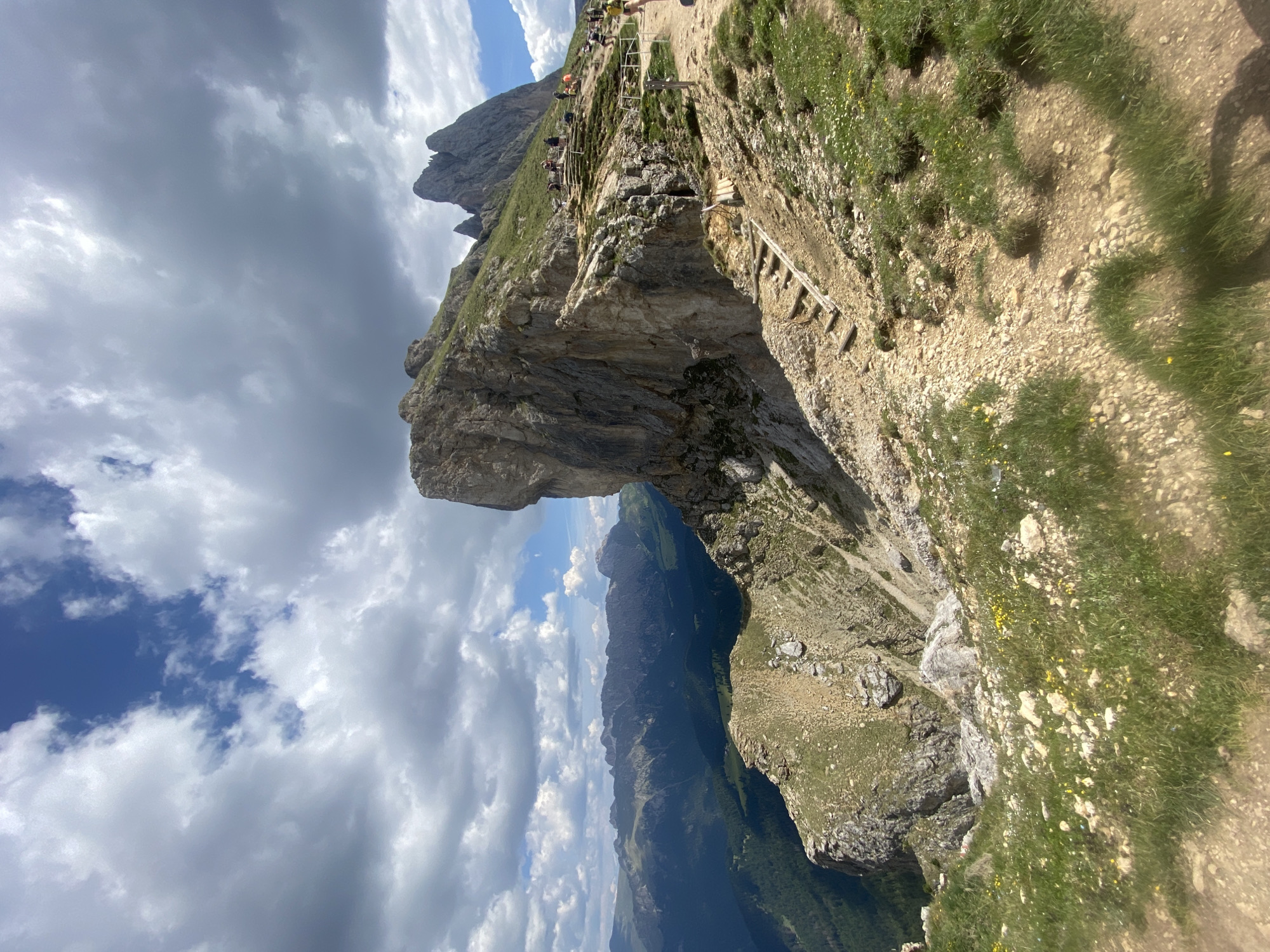 The image depicts a mountainous landscape with a hiking trail. Several people are visible on the trail, which is carved into the side of a steep, rocky cliff. The trail appears to be narrow, with a wooden railing in some sections and a ladder made of wooden planks leading up a steep incline. The terrain is covered with grass and small yellow flowers. In the background, a vast mountain range stretches across the horizon, with deep valleys and lush green slopes. The sky is partly cloudy, with dramatic cumulus clouds casting shadows over the mountains. The scene suggests a sunny day, likely during daytime, suitable for outdoor activities like hiking. The rugged terrain and the presence of hikers indicate a popular spot for mountaineering or trekking in a natural, somewhat remote setting. The overall mood is one of adventure and the beauty of nature. There is no visible text in the image.
