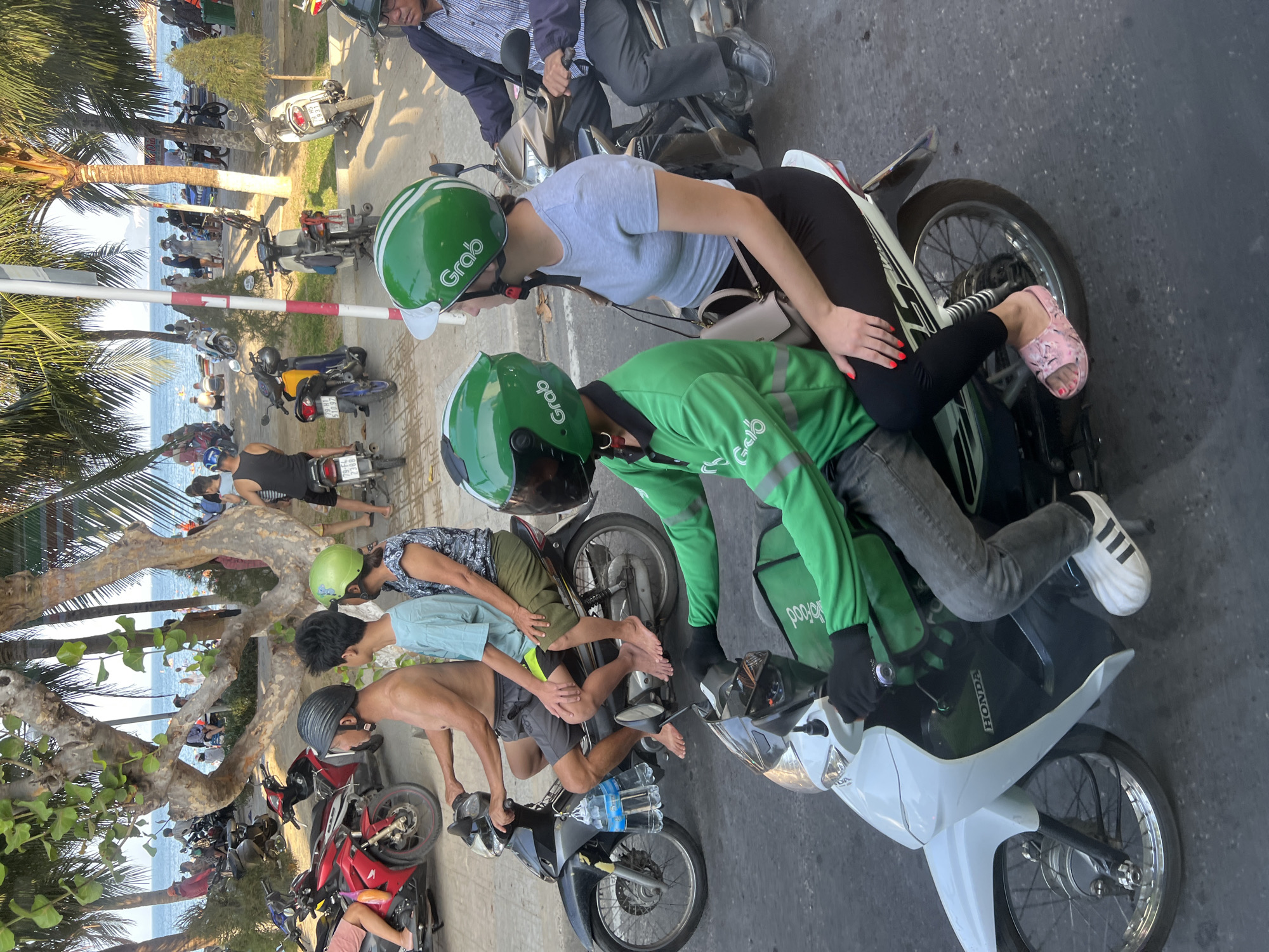 This is a street-level, medium shot of a busy scene on a sunny day in Nha Trang, Vietnam, likely along the beachfront promenade. In the foreground, a person wearing a green Grab uniform and helmet is riding a white scooter, with a passenger wearing a grey t-shirt and black leggings on the back, also wearing a green Grab helmet. Behind them, another scooter carries three passengers: a shirtless man in grey shorts and a black helmet, a young man in a light blue t-shirt and black shorts, and a woman in a patterned top and green shorts. Several other scooters and motorbikes are visible in the background, parked and in motion. The environment includes palm trees, a sidewalk, and the distant ocean with people on the beach and in the water. The overall impression is one of vibrant street life and transportation in a coastal Vietnamese city.