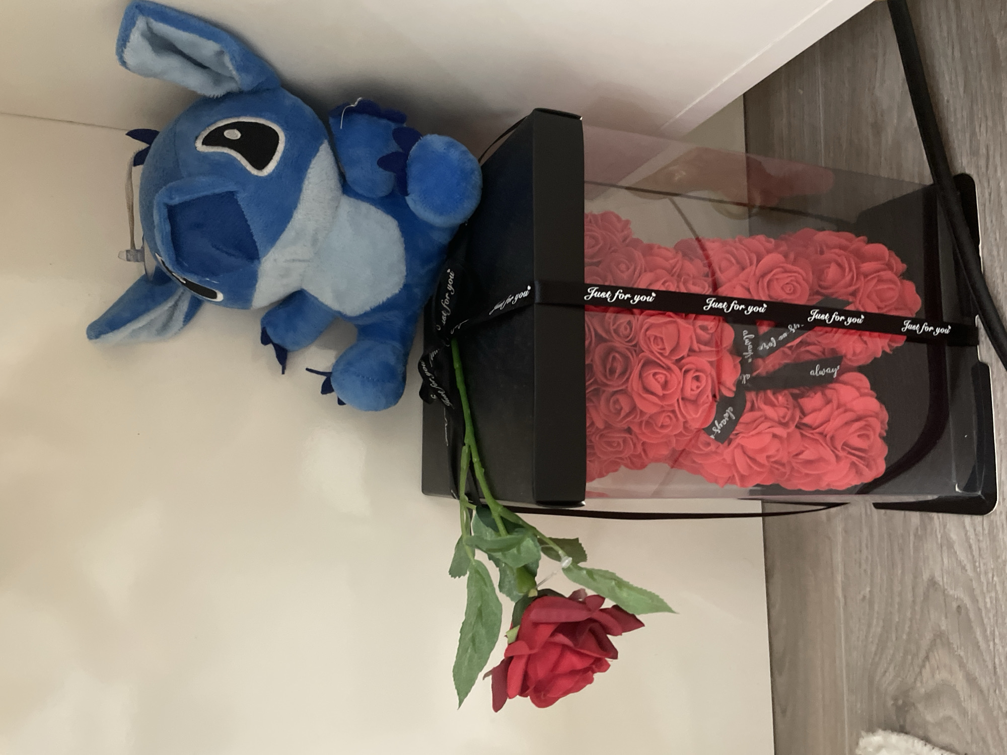 An indoor flat lay image, rotated clockwise, features a blue plush toy, a box of artificial roses, and a single artificial rose. A blue plush toy, resembling Stitch from Lilo & Stitch, occupies the left side, resting on a white surface. Its large black and white eyes and blue fur are clearly visible.
To the right, a black rectangular box with a clear lid contains a heart-shaped arrangement made of numerous small artificial red roses. A black ribbon with white text is wrapped around this box, prominently displaying "Just for you" and "always" multiple times. The OCR also indicates "Bojan Sh." on the ribbon. Hanging down from the side of the box is a single artificial red rose with a green stem and leaves.
The objects are placed on a clean, white surface, possibly a shelf or tabletop. A muted, light-colored wooden surface is visible on the far right, suggesting a wall or floor. The scene is lit with ambient indoor lighting, and no specific time of day is discernible. The arrangement suggests a display of gifts or decorative items, conveying themes of affection or celebration.