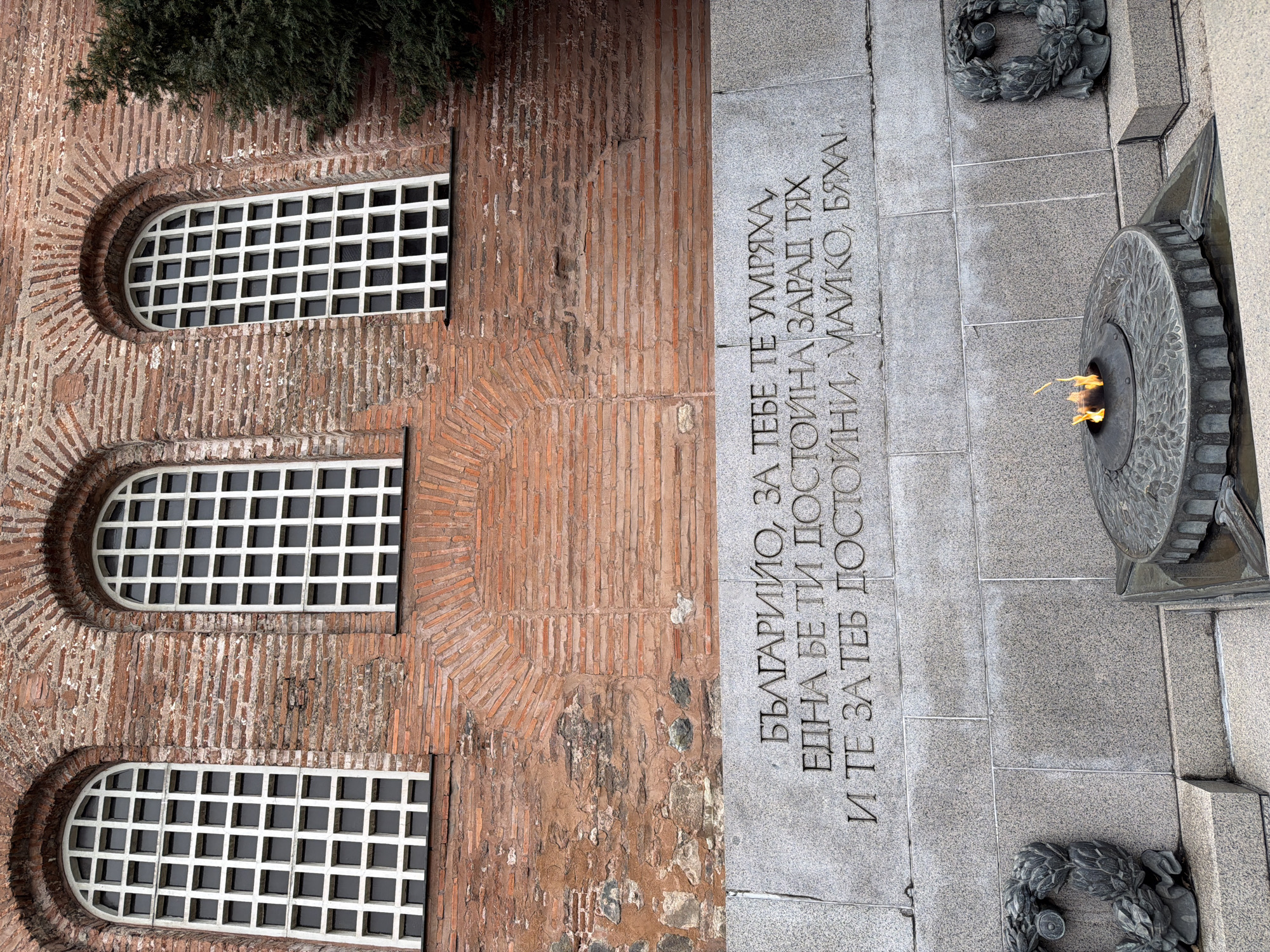 The image shows a section of the Monument to the Unknown Soldier in Sofia, Bulgaria. The left side of the image features a brick wall with three arched windows, each with a grid-like pattern. To the right of the wall, there is a stone slab with an inscription in Bulgarian: "БЪЛГАРИЙО, ЗА ТЕБЕ ТЕ УМРЯХА, ЕДНА БЕ ТИ ДОСТОЙНА ЗАРАД ТЯХ И ТЕ ЗА ТЕБ ДОСТОЙНИ, МАЙКО, БЯХАІ..." ("Bulgaria, they died for you, you were worthy of them, and they were worthy of you, mother!"). Next to the inscription, there is an eternal flame burning in a circular stone structure. At the base of the slab, there are stone wreaths. The scene is captured from a slightly elevated, tilted perspective.