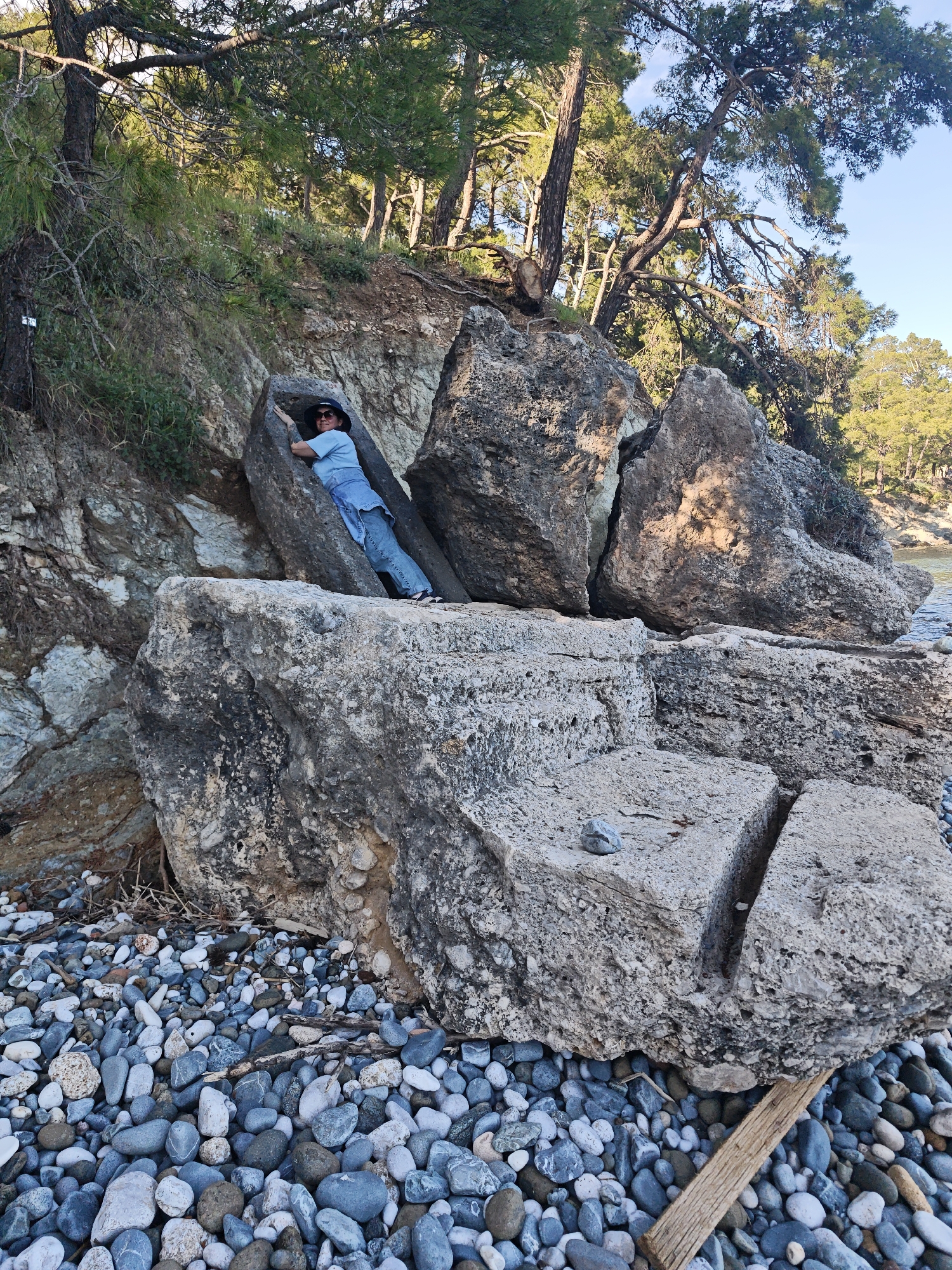 The image captures an outdoor scene at a rocky beach or coastline, likely in Kemer, Turkey, as per the provided context. The ground is covered with numerous small, smooth pebbles in shades of grey, white, and blue.
In the center of the frame, a person is resting or posing among large, weathered rock formations. The individual is a woman wearing a light blue short-sleeved top, blue jeans, a dark blue bucket hat, and sunglasses. She has tattoos visible on her right arm. She is positioned leaning back against one large, dark grey rock, with another rock closing in on her side, creating a natural niche.
Behind the rocks, a steep, rocky slope rises, covered with lush green vegetation, including tall pine trees with characteristic long needles and twisted branches. The trees are bathed in sunlight, suggesting a bright day. The slope leads upwards to a clear sky.
To the right side and in the background, glimpses of calm, blue-green sea or water are visible, indicating the coastal setting. The rocks vary in size and texture, with some showing jagged edges and others being smoother. One large, flat rock in the foreground has a distinct, deep crack running through it, and a small, lighter-colored rock rests on its surface. A single wooden plank is partially visible on the pebbly ground near this rock.
The lighting suggests it is daytime, with clear weather. No visible text is present in the image.