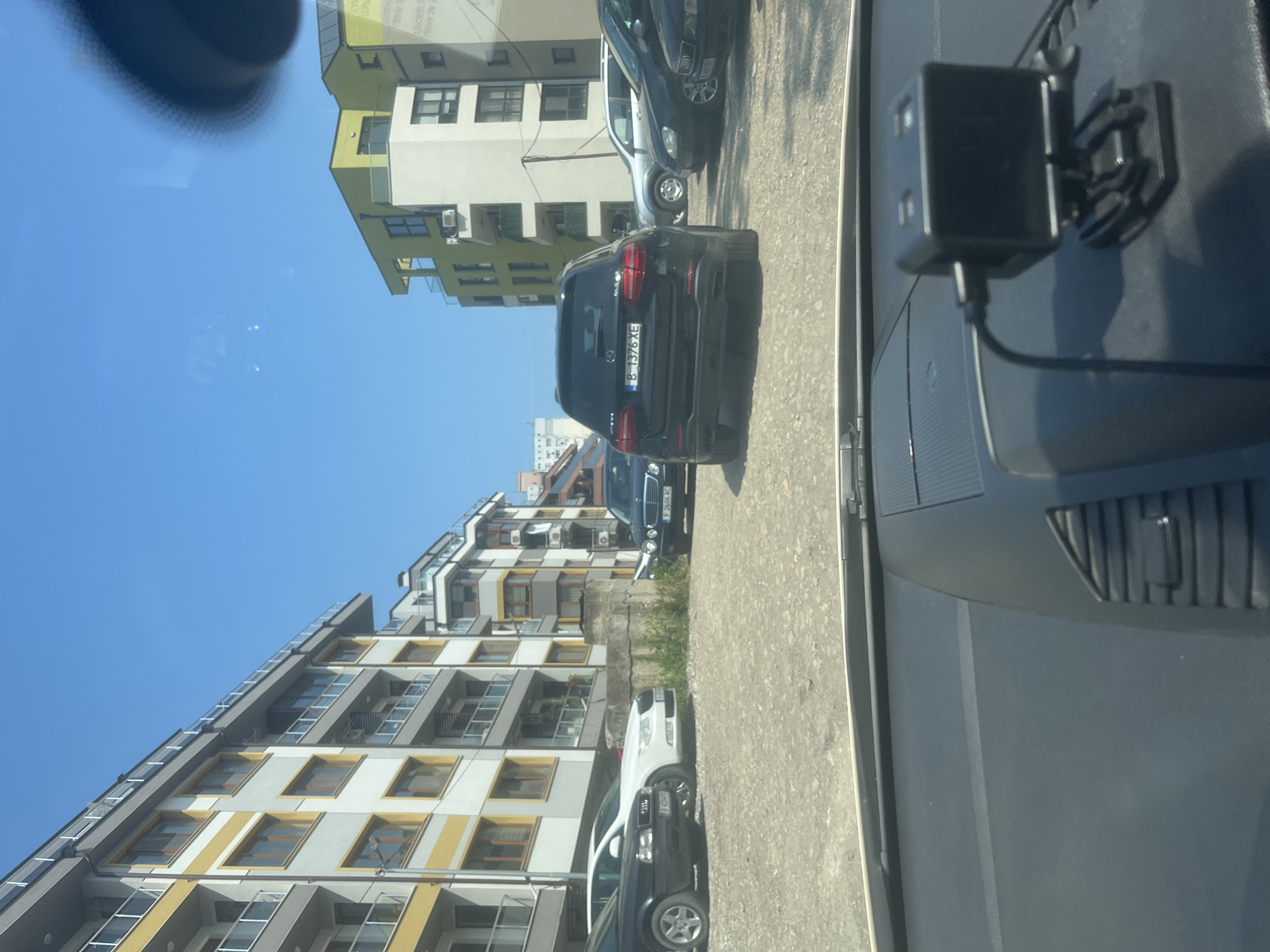 The image captures an urban street scene from the perspective of a vehicle's interior, looking out through the windshield on a bright, sunny day. The view is oriented looking forward and slightly to the right, showing parked cars and apartment buildings under a clear blue sky.
In the foreground, the dark dashboard of the vehicle is visible, with a black dashcam or similar device mounted on its right side. A reflection is noticeable in the upper left portion of the windshield.
The midground features a paved area, likely a parking lot or street, where several vehicles are parked. A black SUV, identified as a Porsche Cayenne by its taillights, is prominently positioned in the center, facing away from the viewer. Its rear license plate, "B 1376 XE," is clearly visible. Further along the street, other cars are parked, including a dark blue sedan with the license plate "Y 2606 KC" and a white SUV.
In the background, modern apartment buildings rise several stories high. These buildings exhibit a contemporary architectural style with various facades, some featuring light-colored walls with yellow or greenish accents, and others with more muted tones. Many windows, balconies, and external air conditioning units are visible on the building exteriors.
The weather is bright and clear, indicated by the vibrant blue sky and strong daylight, suggesting a daytime shot, likely midday or early afternoon. No people are visible in the scene. The overall setting is a bustling urban residential or mixed-use area, consistent with a location in Varna, Bulgaria, despite the "B" on the license plate typically denoting Sofia.