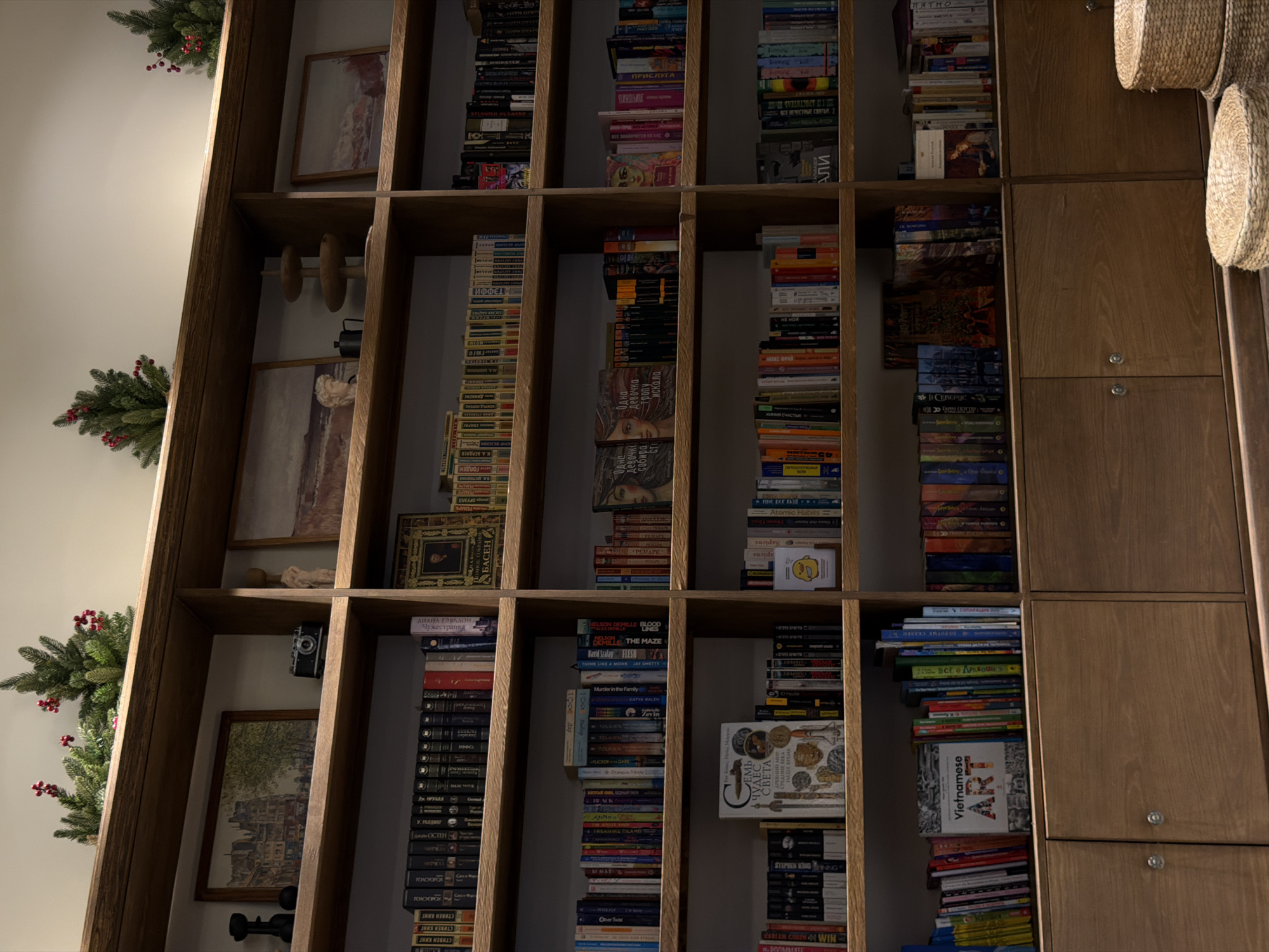 The image shows a tall, wooden bookshelf filled with numerous books, some artwork, and other objects. The bookshelf appears to be built into a corner, with Christmas garland decorating the top and side. The books are arranged haphazardly, covering a wide range of subjects and sizes. Some of the books are visible, including works by Stephen King, Remarque, and others. Other objects on the shelves include picture frames, a vintage camera, and decorative weights. On the right side of the image, some of the bookshelf space is taken up by plain wooden cabinets. The location is Bac Nha Trang Ward, Vietnam.