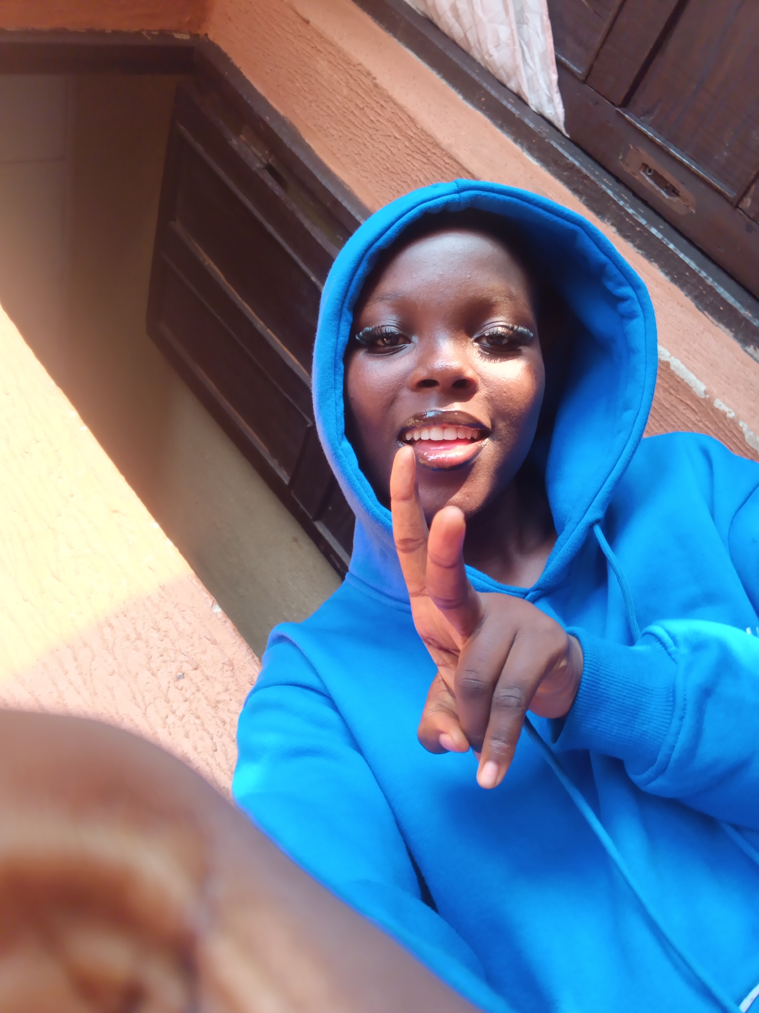 A young Black woman is depicted in a close-up, upward-angle selfie, set outdoors in the city of Onitsha, Nigeria. She is wearing a bright royal blue hooded sweatshirt with the hood pulled over her head. Her face, characterized by a dark complexion, is illuminated by direct sunlight, creating highlights on her forehead and cheekbones. Her eyes feature prominent winged white eyeliner and long dark eyelashes. She presents a playful expression, smiling with her mouth slightly open, revealing her upper teeth and the tip of her tongue. Her left hand (the viewer's right) is raised, with the index finger extended and pointing directly towards the camera; the thumb and other fingers are curled. The background consists of a textured, light orange-brown stucco-like exterior wall and a partially visible dark brown wooden door or window frame with vertical panels and a metallic fixture. The scene is brightly lit, indicating strong outdoor light.