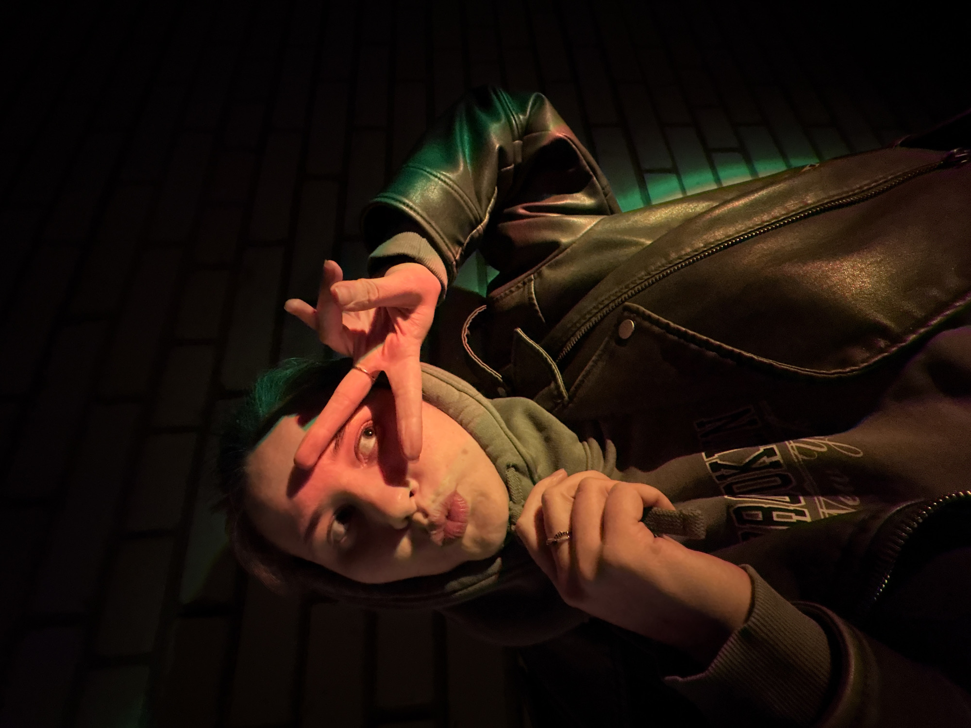 The image depicts a person lying on what appears to be a brick surface, likely outdoors. The person is wearing a hooded sweatshirt and a leather jacket. They are holding up their hand with the middle and index fingers separated, partially covering one eye. The scene is illuminated by a combination of dark and green light, with the brick surface illuminated with green light. The location, Chaikovsky, Russia, is not discernible within the image's frame without additional context or identifying markers. The person's expression is ambiguous.