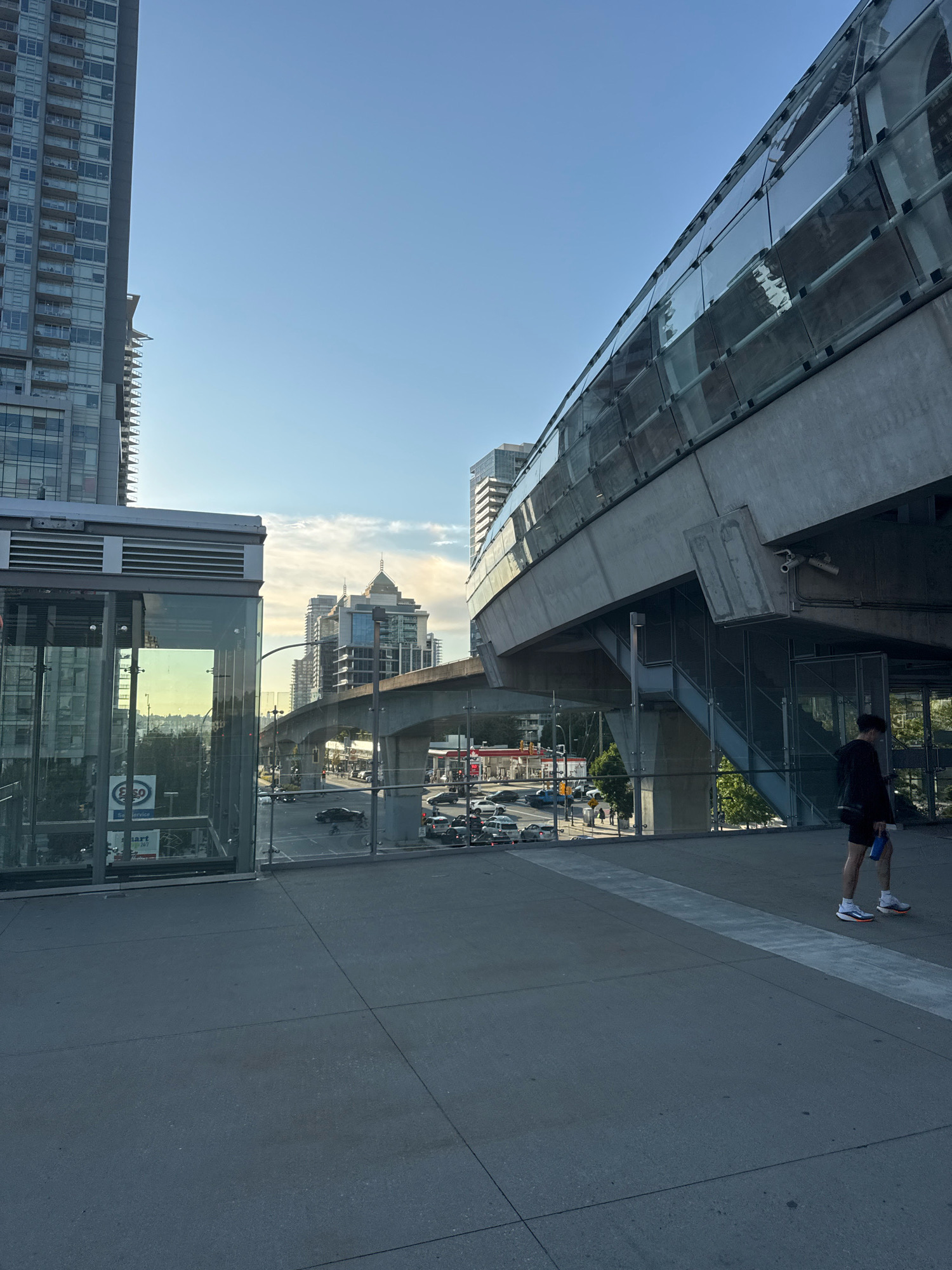 This image depicts an outdoor scene in Burnaby, Canada, likely near a transit station. In the foreground, a person with dark hair, wearing a black t-shirt, black shorts, white socks, and athletic shoes, walks along a paved walkway. They are holding a blue water bottle in their left hand and a black bag over their shoulder. The walkway is part of an elevated structure with a modern design, featuring a concrete base and a glass enclosure along its edge.
To the left of the walkway is a glass-enclosed structure, possibly an elevator or staircase entrance, with visible signage, including an Esso logo. Behind this structure, traffic can be seen on a street below, with cars and a gas station visible in the distance.
Dominating the background are several high-rise buildings, characteristic of an urban environment. An elevated transit line, supported by concrete pillars, curves through the scene. The sky is clear and blue with some wispy clouds, indicating daytime. The overall impression is a contemporary urban landscape with a focus on public transit infrastructure and surrounding city architecture.