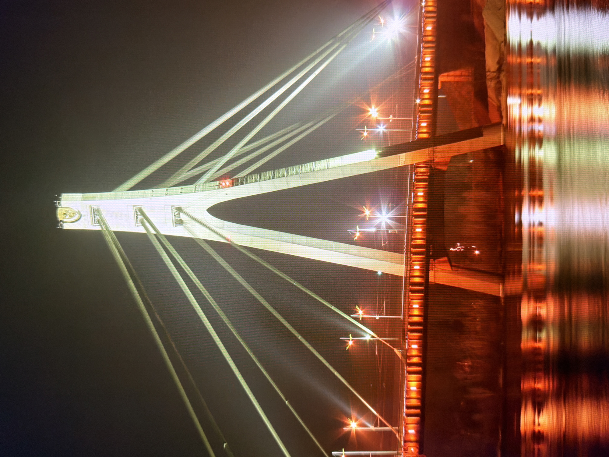 The image depicts the North Bridge (Pivnichnyi Mist) in Kyiv, Ukraine, at night. The bridge is a cable-stayed bridge illuminated by numerous lights. The main support tower, a tall, white concrete structure, is prominently featured, with cables extending outwards to support the bridge deck. The deck itself is lined with orange lights, creating a warm glow. The water below the bridge reflects the lights, creating a shimmering effect. Streetlights with starburst patterns are visible along the bridge. The background is dark, suggesting a night scene in an urban environment. The overall impression is one of a modern, illuminated city infrastructure.