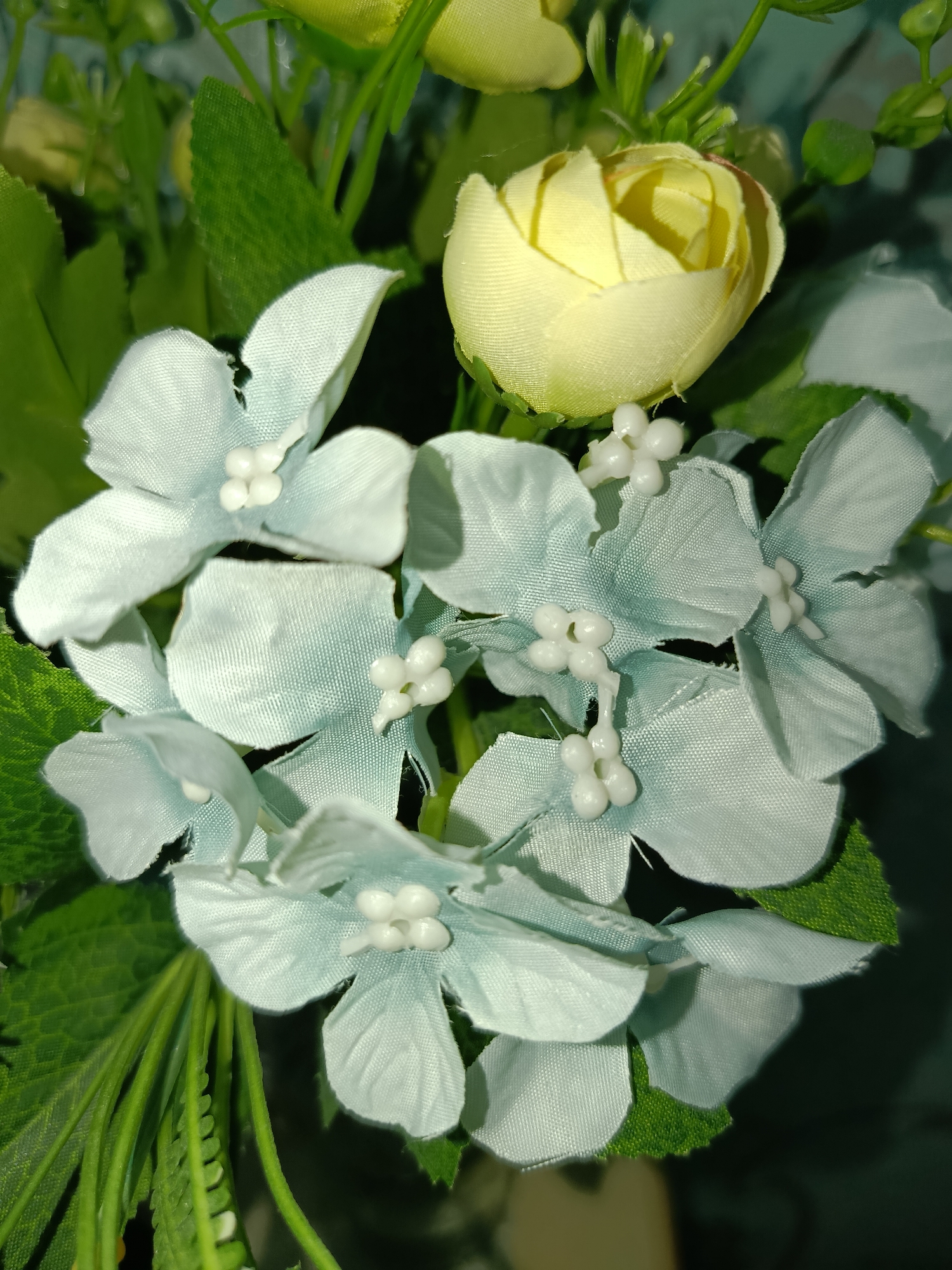 A close-up, overhead shot captures a bouquet of artificial flowers. The primary focus is on several light blue, five-petaled flowers arranged in a cluster. Each of these flowers has small, white stamen-like protrusions in the center. Partially visible above and to the right of the blue flowers is a single, tightly closed yellow rosebud. Lush green artificial leaves and stems fill the background, creating a soft focus. The lighting appears to be artificial, casting shadows and highlighting the textures of the fabric flowers. The image does not contain any identifiable landmarks or elements that would suggest a specific location within Tashkent, Uzbekistan, or any other city.