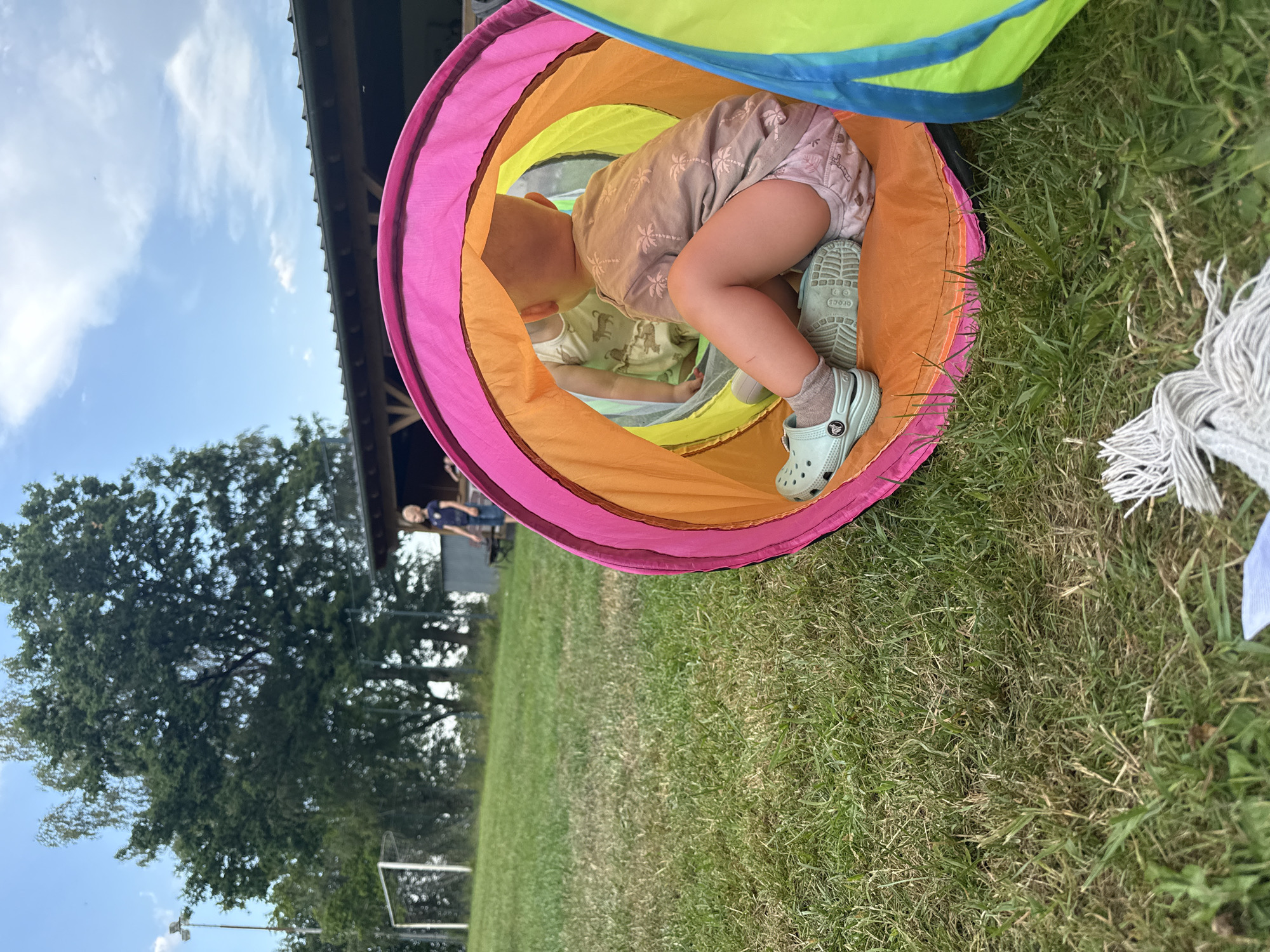 In Lumda, Germany, a young child sits inside a colorful play tunnel on a grassy field. The tunnel, which appears to be made of fabric with pink, orange, and yellow sections, is positioned on the ground. The child, seen from behind, is wearing a light-colored t-shirt with a palm tree pattern and light-colored shorts with a subtle print. They have on light blue Crocs and grey socks. Another child is partially visible inside the tunnel, facing away from the camera. In the background, there is a soccer goal, trees, and a covered structure with a roof. The sky above is blue with scattered clouds.