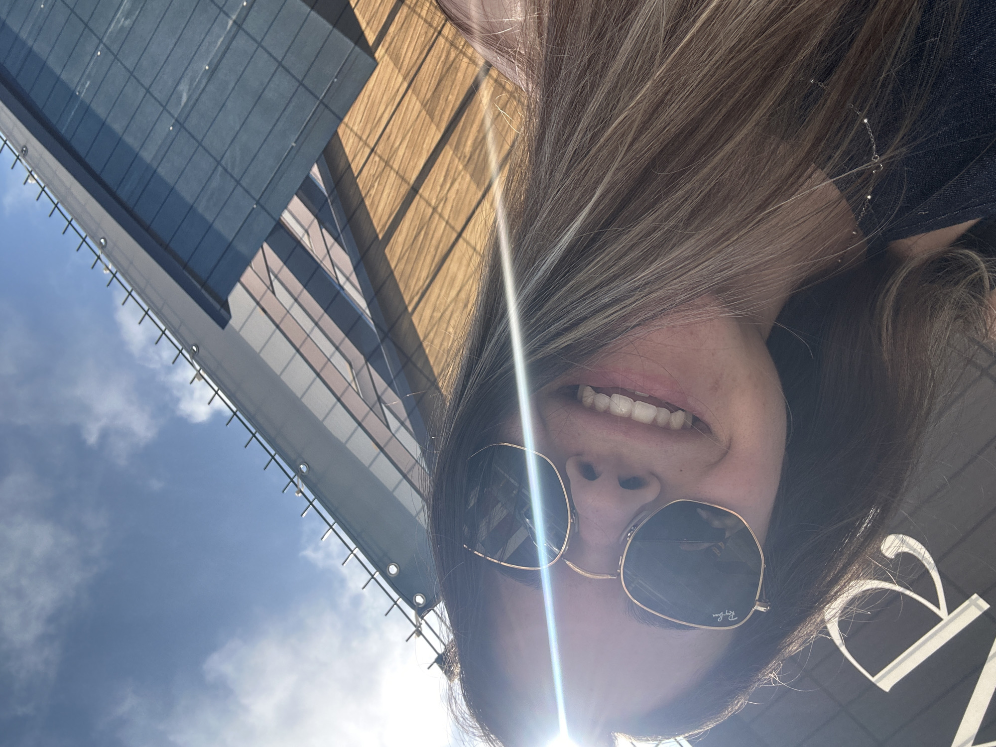 An overhead, low-angle shot captures a person, likely a woman, looking upwards with a smiling expression, revealing white teeth. Her long, light brown hair with blonde highlights frames her face and falls around her. She wears round, gold-rimmed sunglasses, with the brand "Ray-Ban" visible on the right lens. A bright sun flare emanates from the lower right of the image, indicating direct sunlight and a sunny day.
In the background, a modern building with a facade featuring dark blue rectangular panels and light brown/wooden sections is visible. The architecture suggests a contemporary urban environment. Part of a blue sky with scattered white clouds is also present. A white banner-like structure with horizontal bars runs along the top edge of the frame, possibly part of the building's exterior or a nearby scaffold. To the right of the person's head, a partial white letter "B" is seen against a dark background, suggesting a sign or logo. The overall scene conveys a cheerful mood under bright, sunny conditions in Sofia, Bulgaria.