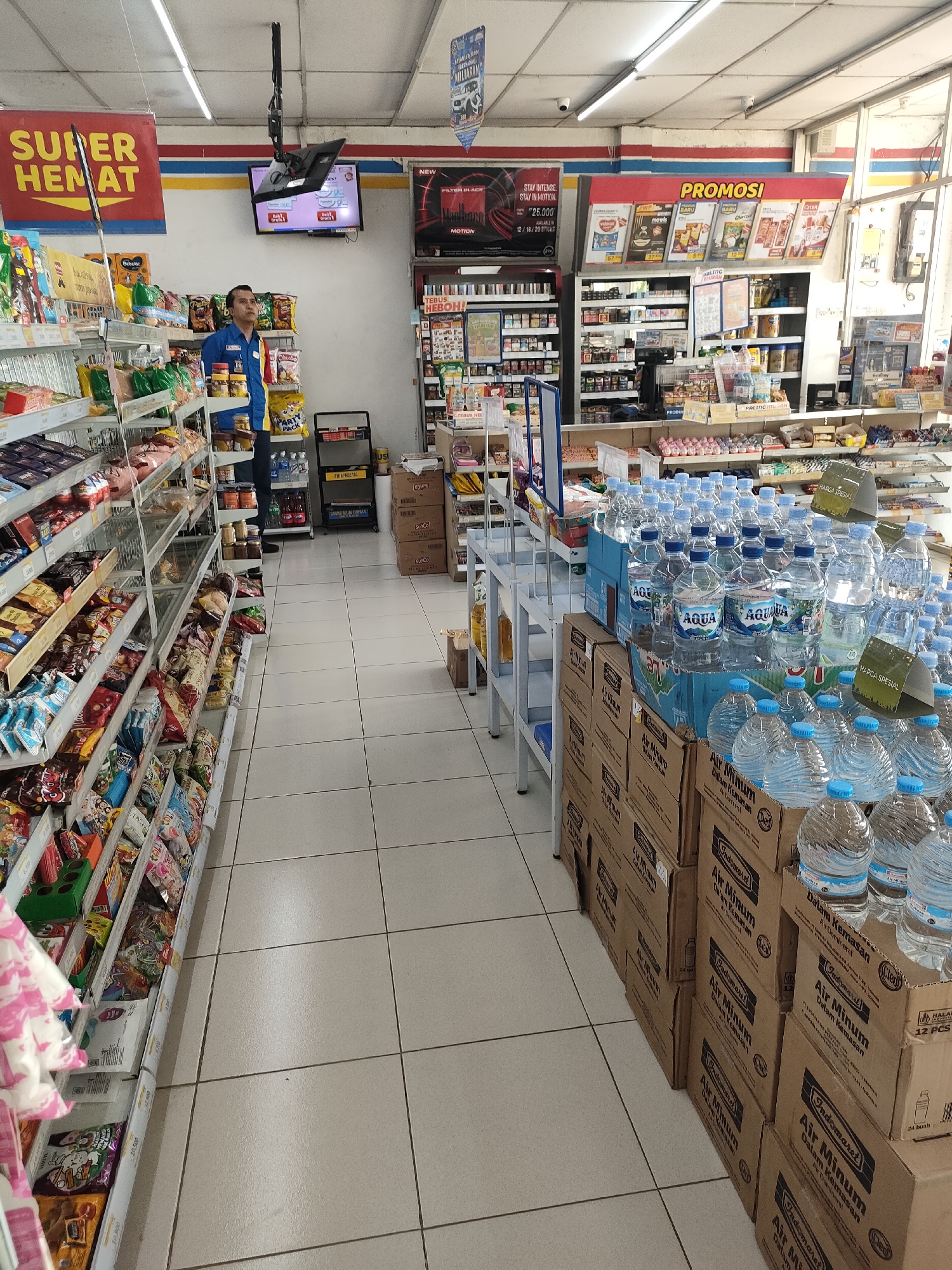 This is a medium-shot, eye-level photograph of the interior of a convenience store. The store appears to be a branch of Indomaret, a popular convenience store chain in Indonesia, as indicated by the branding on product boxes and promotional materials. The store is well-lit with fluorescent ceiling lights and features rows of shelves stocked with various consumer goods, including snacks, bottled beverages, and other packaged items.
In the foreground, on the right side of the image, a large display of bottled water, predominantly Aqua brand, is arranged on top of cardboard boxes. The boxes themselves are branded with "Indomaret Air Minum Dalam Kemasan." Several of these water bottles have green "HARGA SPESIAL" (Special Price) tags attached to them.
The left side of the image shows aisles of shelves filled with a variety of packaged snacks and other products. The shelves are organized and stocked, with clear price labels visible on some items.
In the background, a male store employee wearing a blue uniform with a yellow collar and a blue name tag is standing near a shelving unit. He is looking towards the ceiling. Above him, a flat-screen television is mounted, displaying what appears to be a menu or promotional content. Further in the background, more shelves and promotional displays are visible, including a "PROMOSI" (Promotion) banner. The flooring is tiled in a light beige color. The overall environment suggests a typical, organized retail space. The location is confirmed to be Curahdringu, Indonesia based on the context provided.