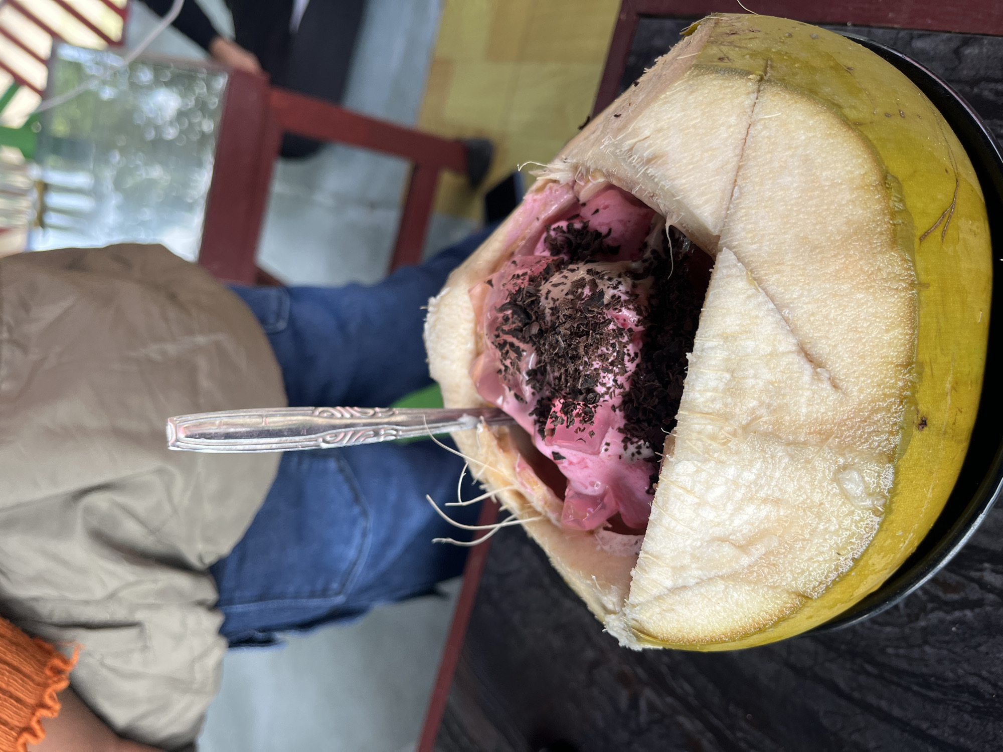 The image shows a half-cut young coconut filled with pink ice cream and chocolate shavings, sitting on a black plate. A silver spoon is inserted into the ice cream. The coconut rests on a dark wooden surface, possibly a table. An individual is seated at the table, wearing blue jeans, a tan shirt, and a glimpse of an orange sleeve. Behind the table, parts of a red wooden chair and other people are visible. The image was likely taken in Lhokseumawe, Indonesia, given the query context. The focus is on the coconut dessert and the person consuming it in an outdoor setting.
