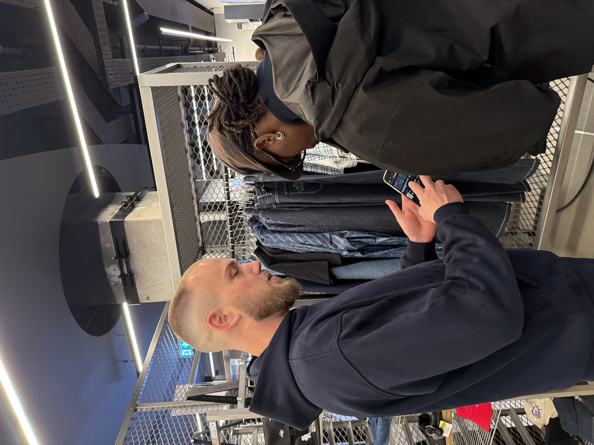 The image shows two individuals in what appears to be a retail environment.
One person, a man with a shaved head and beard, is wearing a navy blue hoodie. He is looking at a mobile phone held by the other individual.
The second person, also a man, is wearing a brown cap, dreadlocks, and glasses. He is also wearing a black jacket and holding a smartphone.
They both stand near clothing racks filled with folded jeans and shirts behind a wire mesh rack. The environment includes industrial-style lighting and metal shelving and racks.
While I can see that the racks are arranged as one would find clothing in a retail shop, I cannot confirm it is located in Paris, France.
