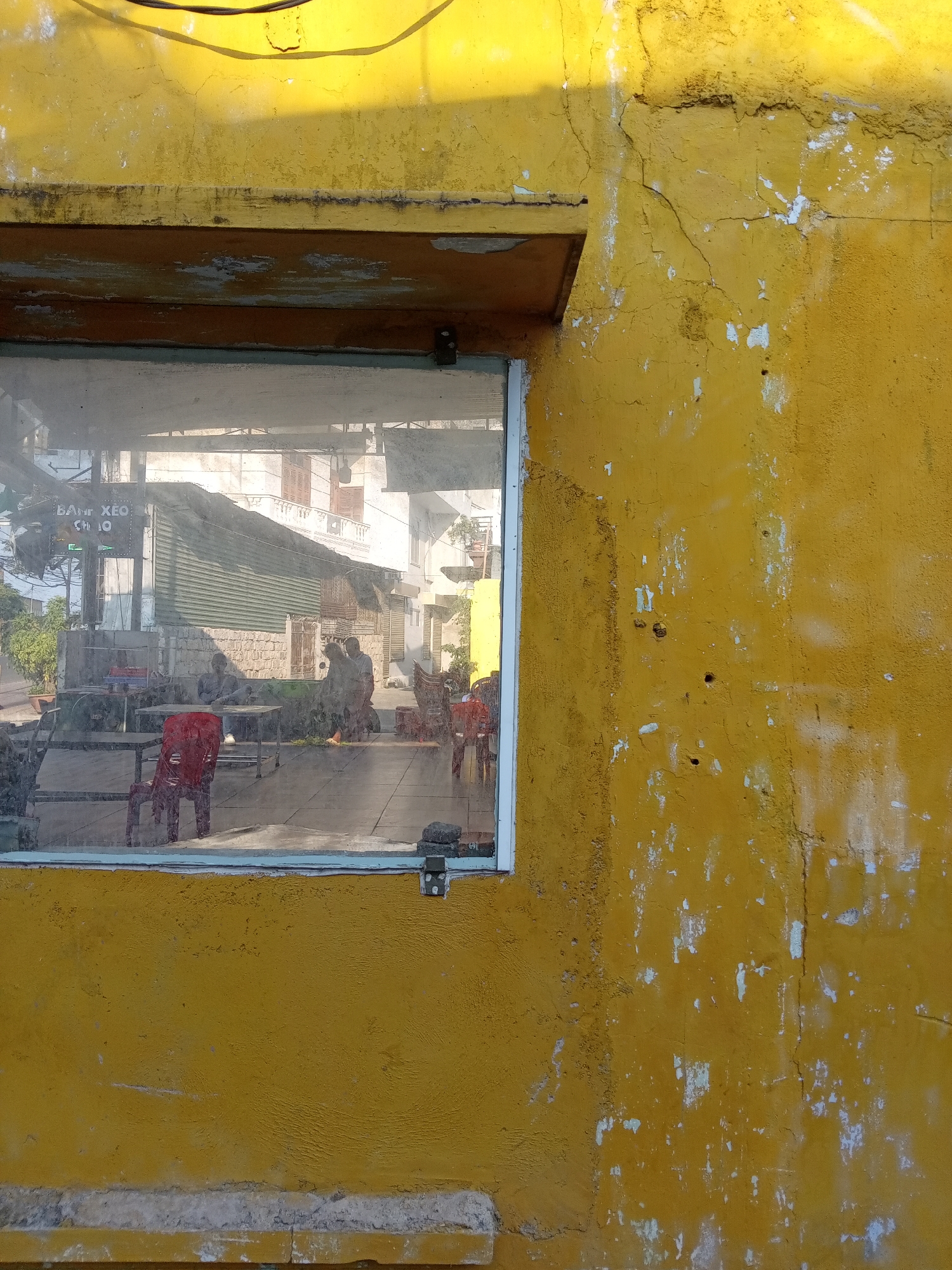 The image shows a yellow concrete building with a window reflecting a street scene in Bắc Nha Trang, Vietnam. The building's exterior is weathered, with chipped paint and visible cracks. The window reflects a building across the street with an awning reading "BANH XEO CHA O." People are seated at tables on the outdoor patio. Red plastic chairs are visible. The overall light in the scene is bright, with harsh shadows. The image is taken from an eye-level perspective.