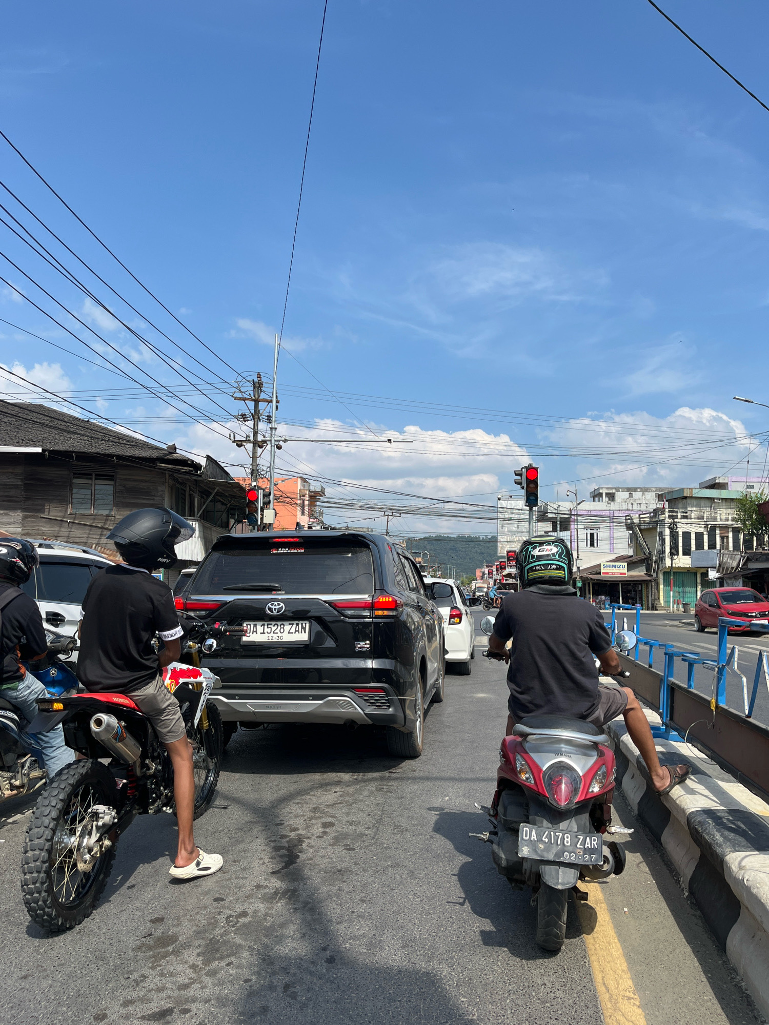 The image depicts a street scene in Bersujud, Indonesia, with several vehicles stopped at a traffic light. The sky is blue with scattered clouds. In the foreground, two motorcycles are visible. The motorcycle on the left is a dirt bike, ridden by a person wearing a black helmet, black t-shirt, and gray shorts. The motorcycle on the right is a scooter, ridden by a person wearing a helmet with green and black accents, a black t-shirt, and gray shorts. Both riders are facing forward, indicating they are waiting at the traffic light.
Behind the motorcycles, a black SUV is stopped. The license plate reads "DA 1528 ZAN". Further ahead, other vehicles, including a white car and a red car, are also visible in traffic. Buildings line the street, with some appearing older and made of wood, while others are more modern. Power lines are strung overhead. The traffic light is red, indicating that the vehicles are stopped. The road surface is asphalt with a yellow line marking a lane. A concrete barrier with blue fencing is on the right side of the road.