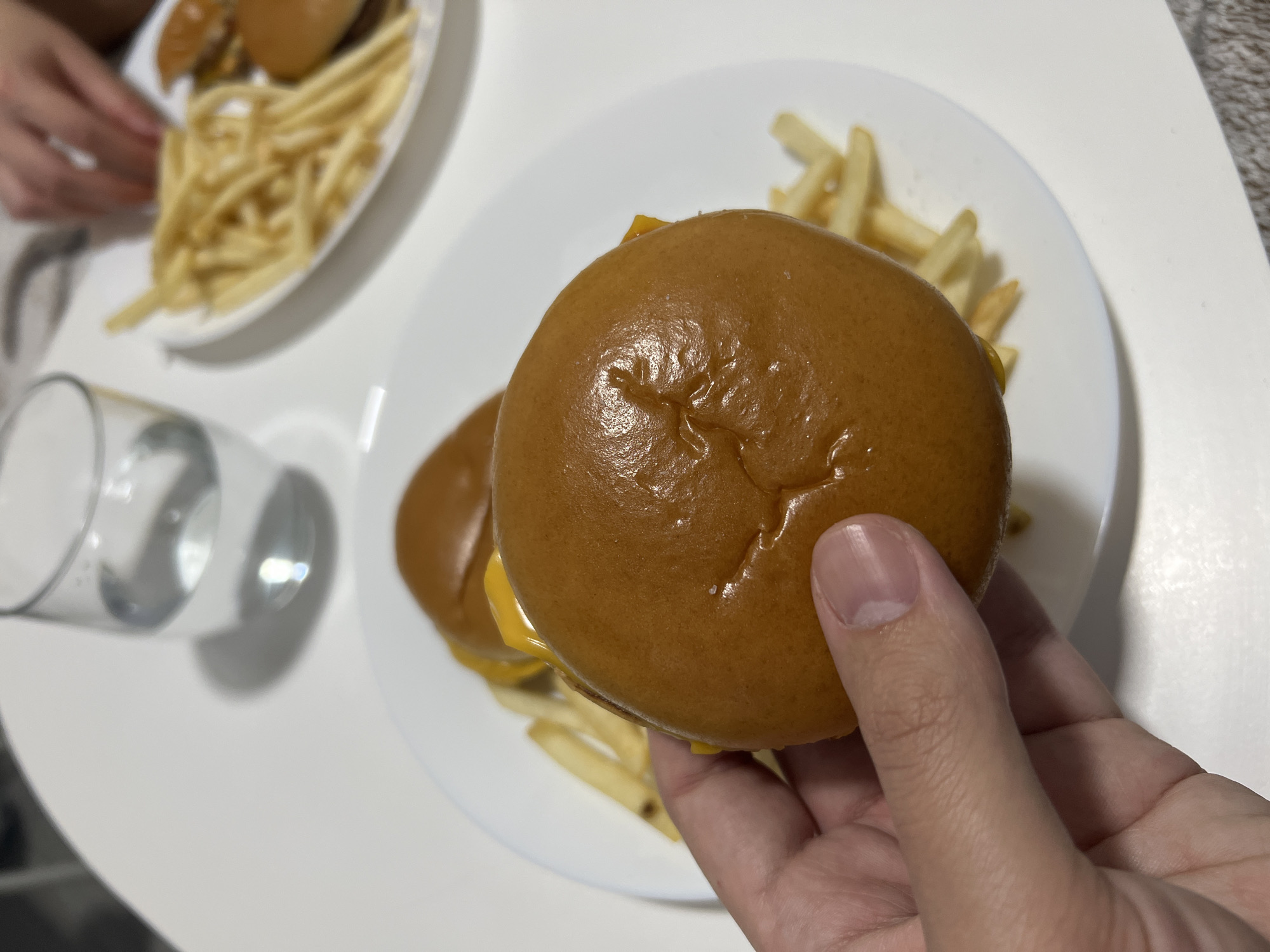 A person is holding a hamburger in their right hand. The hamburger has a glazed bun, and cheese is visible between the bun and the filling. The hamburger rests on a white plate with french fries. Another plate with fries and burgers is in the background. A partially filled glass of water sits on a white table. The location is unidentified.