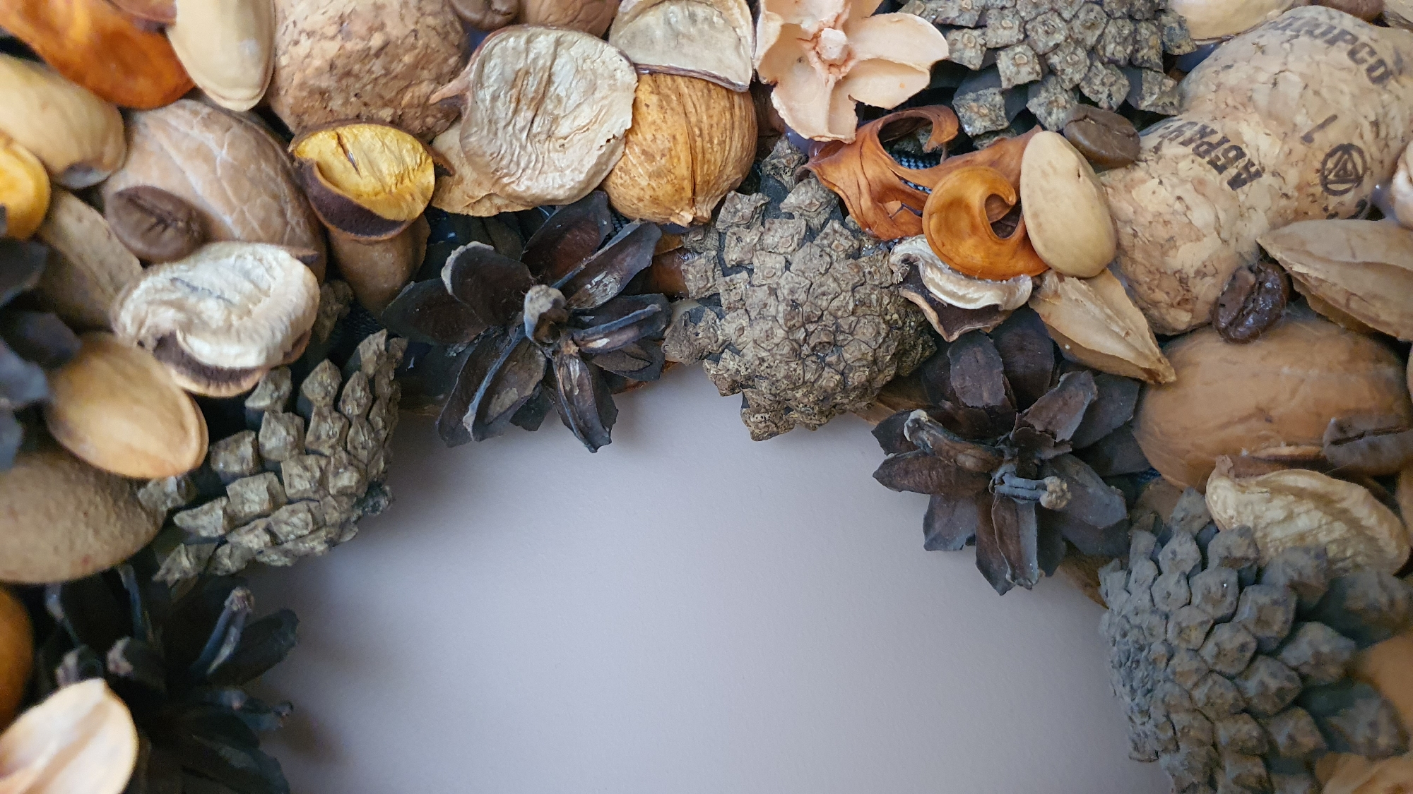 The image is a close-up, vertical shot of a decorative arrangement of natural elements, likely intended as a wreath or a decorative centerpiece. The items are primarily dried botanicals, including pinecones of various sizes and textures, nuts (some shelled, some unshelled), dried seed pods, and what appear to be dried flower petals or leaves. The color palette is predominantly earthy tones, featuring shades of brown, beige, cream, and some darker, muted blues and grays from the pinecones. The background is a plain, light-colored surface, possibly a wall or fabric, which is out of focus. The arrangement is dense and textured, with the different elements overlapping and creating a layered effect. Some of the items, like the pinecones, have intricate, scaly surfaces, while others, such as the nuts and seed pods, are smoother or more flattened. There is a hint of texture on one of the corks that reads "АБРА" and "ОРФ". The lighting appears to be soft and diffused, highlighting the textures of the natural materials. The image does not contain any discernible elements that would suggest a specific location within the city of Krasnoyarsk, Russia, or any human presence or activity.