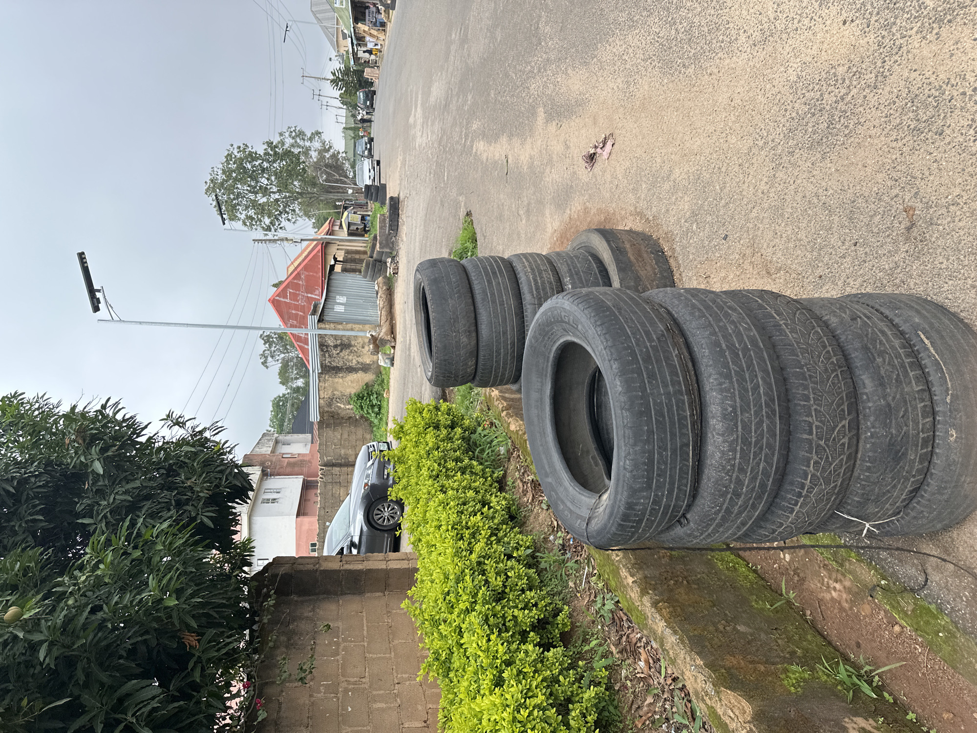 The image captures a roadside scene with stacks of used tires placed on the side of a paved road. In the foreground, a pile of six tires is stacked vertically. To the left of the tires, a dense green hedge runs along a concrete curb, separating it from a brick wall. Beyond the wall, a dark-colored sedan is partially visible. In the background, the road continues, with several cars and people visible further down. Buildings and trees line the street, and utility poles with wires are present overhead. The sky is overcast. The overall environment suggests a street in a town or city.