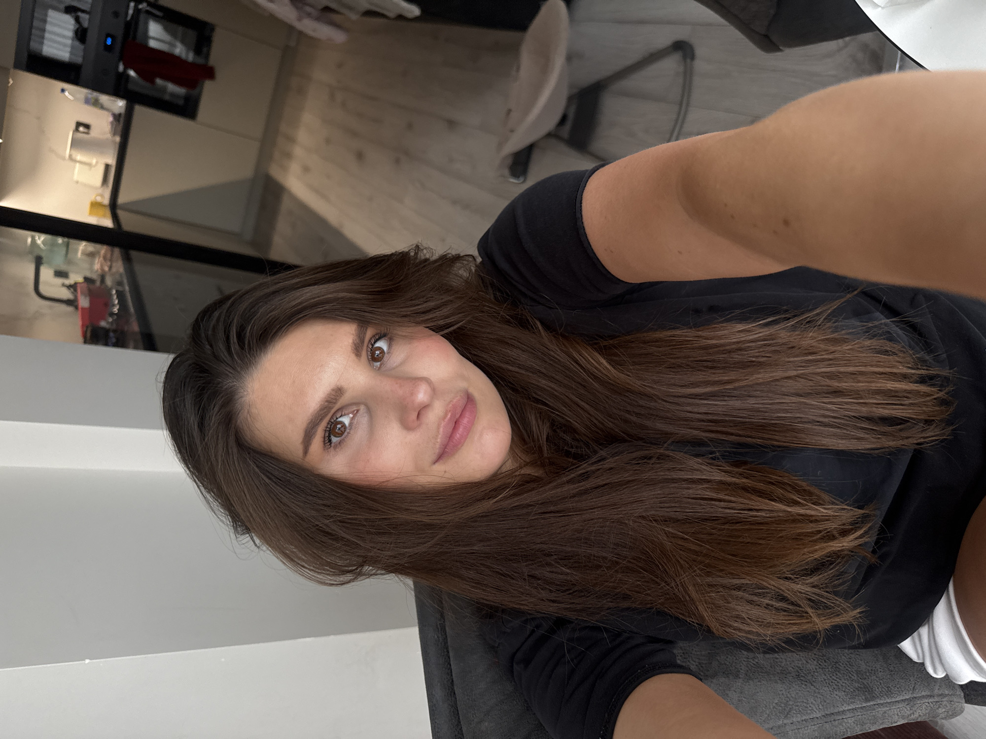 The image presents a selfie of an adult female, reportedly located in Sofiivska Borshchahivka, Ukraine. She is depicted from the chest up, with her right arm extended to hold the camera, suggesting a seated position against a dark grey upholstered surface, likely a chair or sofa. Her long, straight dark brown hair is parted near the center and falls over her shoulders. She has brown eyes and a fair complexion, with light makeup accentuating her eyes and lips, and a subtle smile. She wears a black short-sleeved t-shirt, with a sliver of white fabric, possibly shorts, visible at the bottom edge of the frame.
The background reveals a domestic interior. To the woman's upper right (viewer's left), a light-colored, plain wall is visible, showing a distinct corner. To her left (viewer's right), a modern kitchen area is in focus. The kitchen features light wood laminate or plank flooring, cream-colored base cabinets, and dark countertops. Various items are visible on the counter, including a white electric kettle, a yellow mug, a clear water bottle, and a dark faucet over a sink area. A dark built-in oven or stove unit is positioned on the right side of the kitchen, with a red cloth draped over its handle and a blue light illuminated on its display panel. Under-cabinet lighting provides illumination to the counter space. In the lower right corner of the background, on the wooden floor, a light-colored, modern-design baby bouncer or infant chair with a minimalist frame is present.