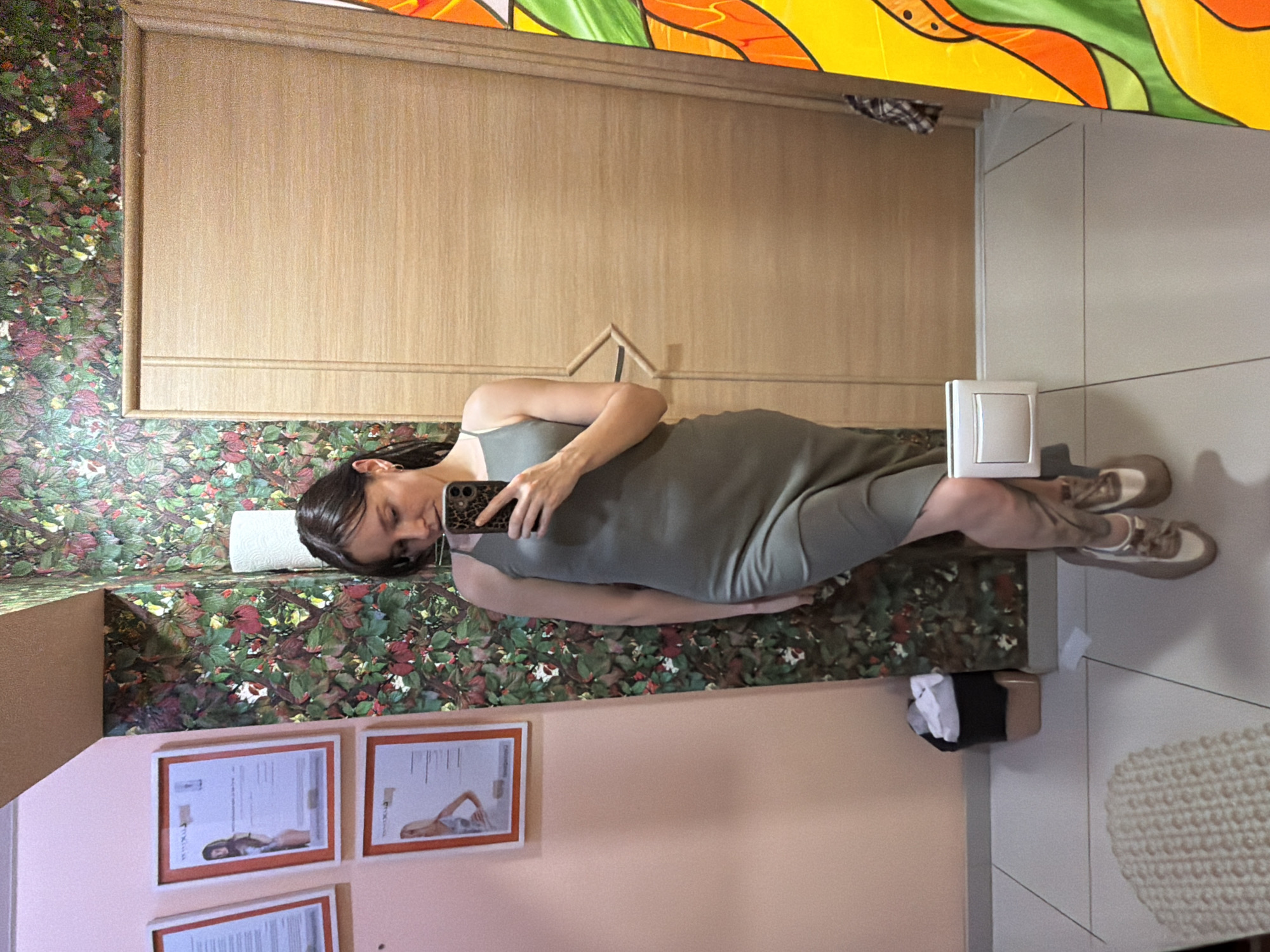 A woman is taking a mirror selfie in a room. She is wearing a grey dress and sneakers. The room has floral wallpaper and wooden paneling. There are framed pictures on the wall. A stained-glass window is visible in the upper part of the image. The woman's expression is neutral. The scene appears to be indoors, possibly a bathroom or changing room.