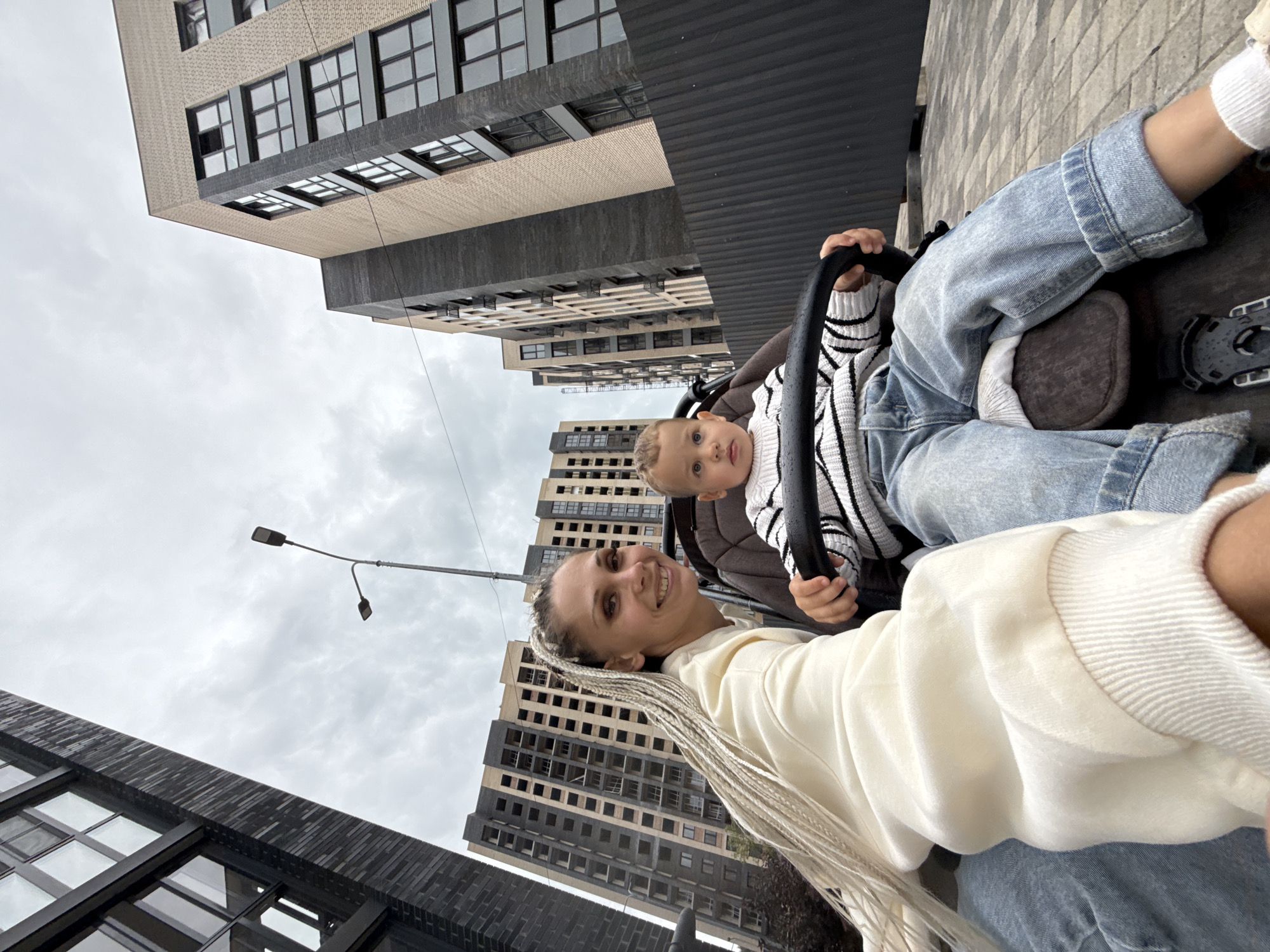 In Rusanovo, Russia, a mother with blonde braided hair smiles at the camera while her young child sits in a stroller. The child, wearing a striped sweater and blue jeans, holds onto the stroller's handlebar. Behind them, modern apartment buildings with dark and beige facades rise against a cloudy sky. A black metal fence separates the foreground from the buildings. The scene appears to be a street or pedestrian area with cobblestone paving.