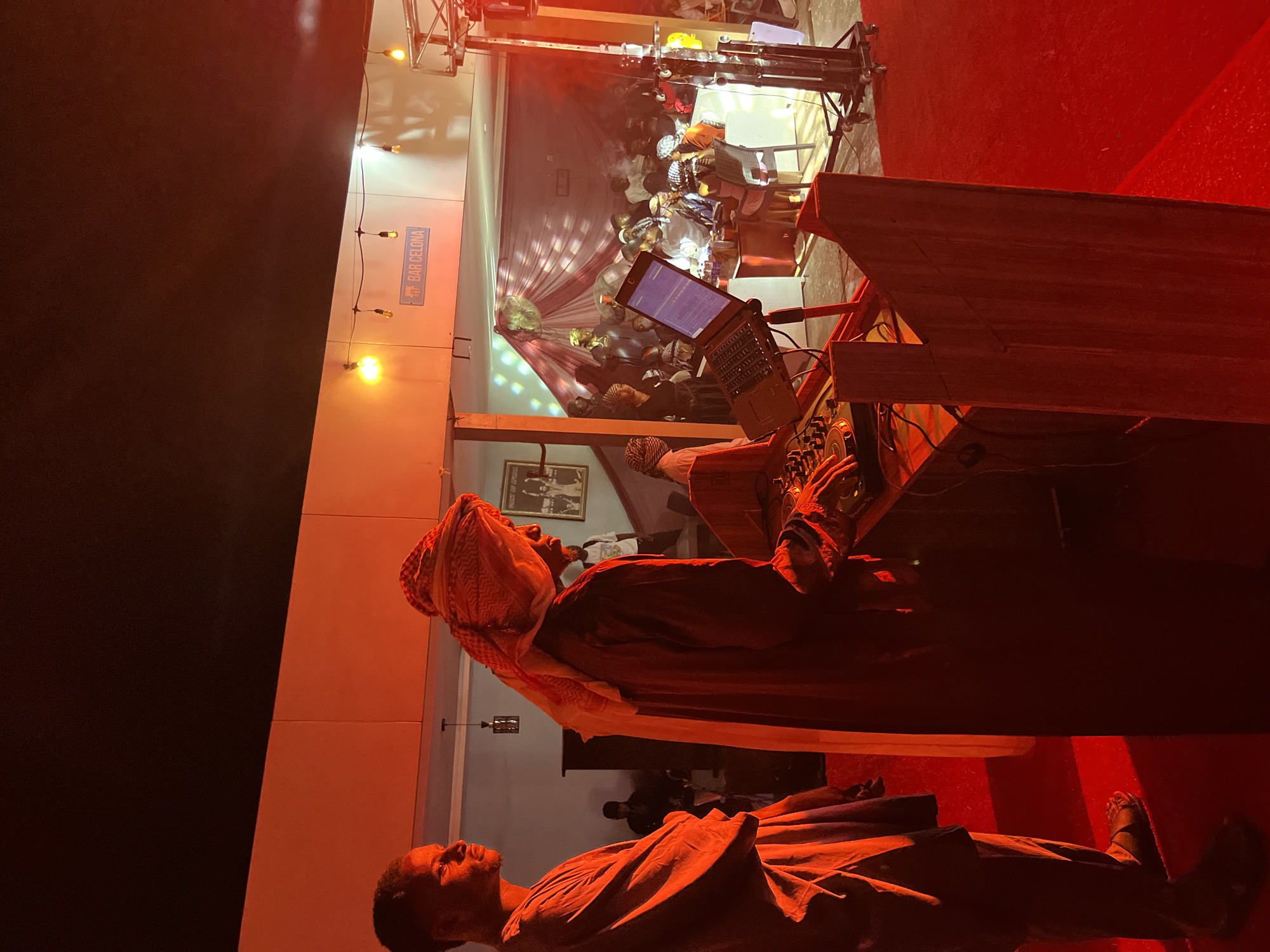 The image depicts a predominantly red-lit indoor event space. In the foreground, two adult males stand on a red carpet. The male on the left, shown in profile facing right, wears a dark, long-sleeved tunic-style garment and dark open-toed sandals. The male on the right, also with a dark complexion, wears a patterned red and white ghutra-style head covering and a dark, flowing robe. He stands behind a dark wooden DJ console, actively manipulating its controls with both hands. An open laptop, displaying what appears to be audio waveform software, rests on a stand to his right.
Above the main activity area, a light-colored horizontal architectural beam spans the scene. Affixed to this beam are string lights with exposed bulbs and a blue rectangular sign with white text that reads "BAR CELONA" with a small emblem above "BAR".
In the background, beyond the DJ setup, an elevated or recessed area is visible, populated by approximately 15 to 20 individuals, both standing and seated at tables. Many of these individuals wear various head coverings and traditional-style garments. This area is decorated with red and white fabric drapes and appears hazy, likely due to a fog machine. Reflections from a disco ball or similar lighting effect are visible on the back wall, along with a wall-mounted fan and a framed picture. On the far right, metal stage trussing or lighting equipment stands vertically. The overall environment suggests a social gathering or performance event.