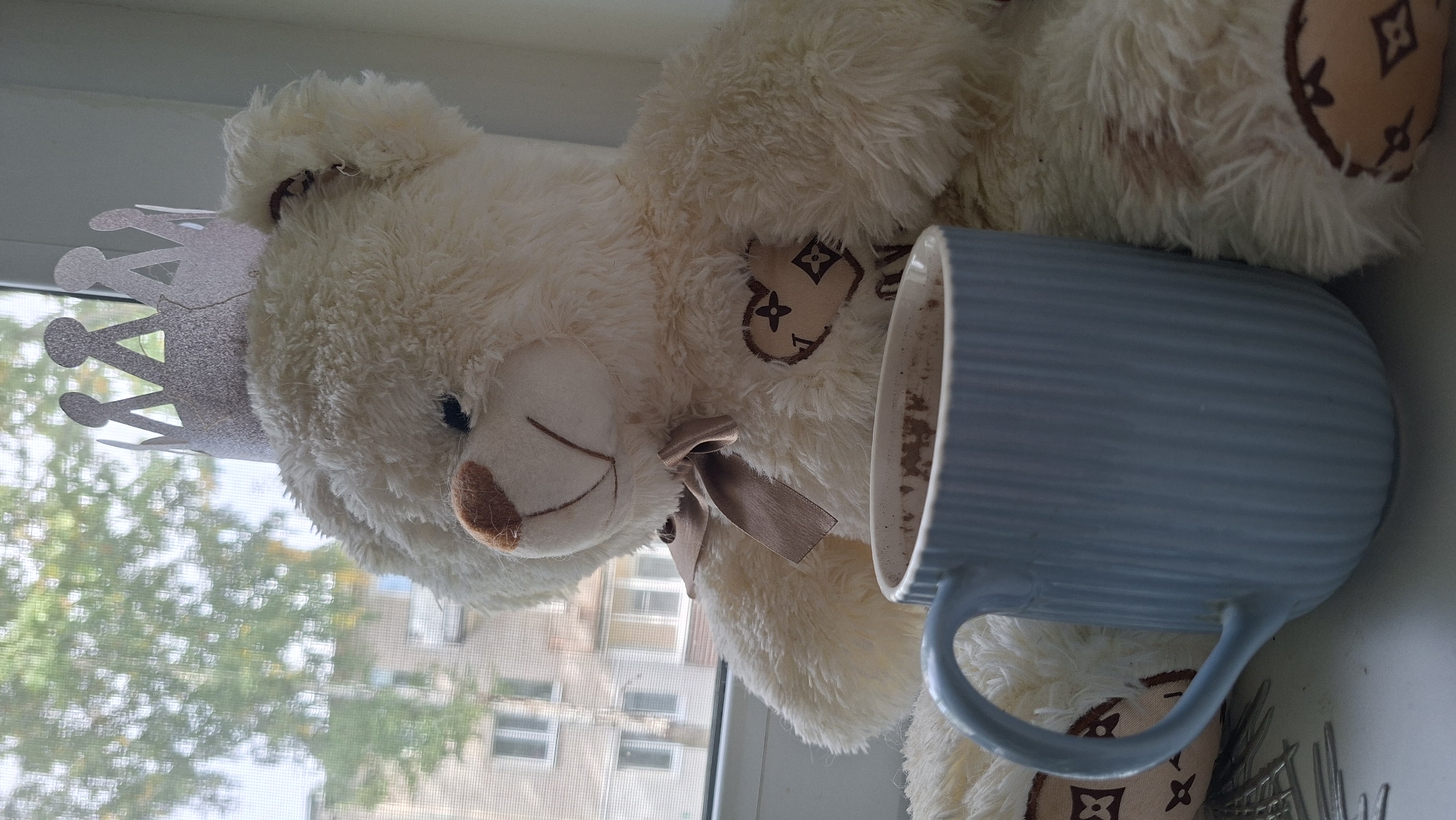 A cream-colored teddy bear wearing a silver glittery paper crown sits on a windowsill. The teddy bear has a brown nose and a brown ribbon tied in a bow around its neck. A light blue ribbed mug sits in front of the teddy bear. The mug contains a dark liquid, possibly hot chocolate. In the background, through the window, is a blurry view of trees and a multi-story building, suggesting an urban or suburban setting. The overall impression is a cozy, indoor scene with a whimsical touch.