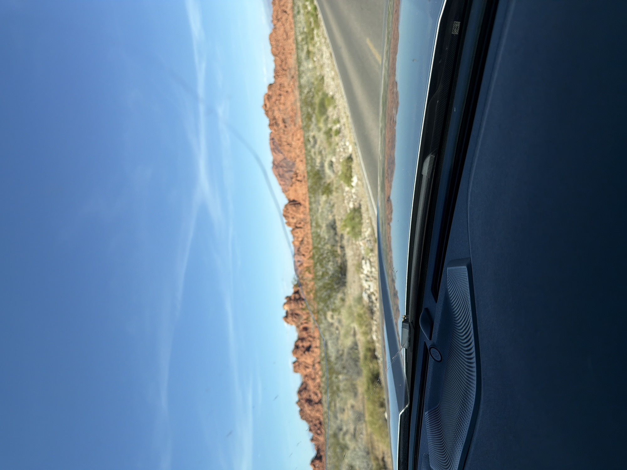 The image depicts a view through the windshield of a vehicle, likely a car. The exterior scene is of a landscape with red rock formations, sparse green vegetation, and a road. The sky is clear blue with some wispy clouds. The location appears to be in a desert environment, reminiscent of the southwestern United States. The road is asphalt, with a yellow line.
Inside the vehicle, part of the dashboard is visible. A black speaker is seen in the interior.
Unknown, United States of America is used as a stand-in for the specific location.