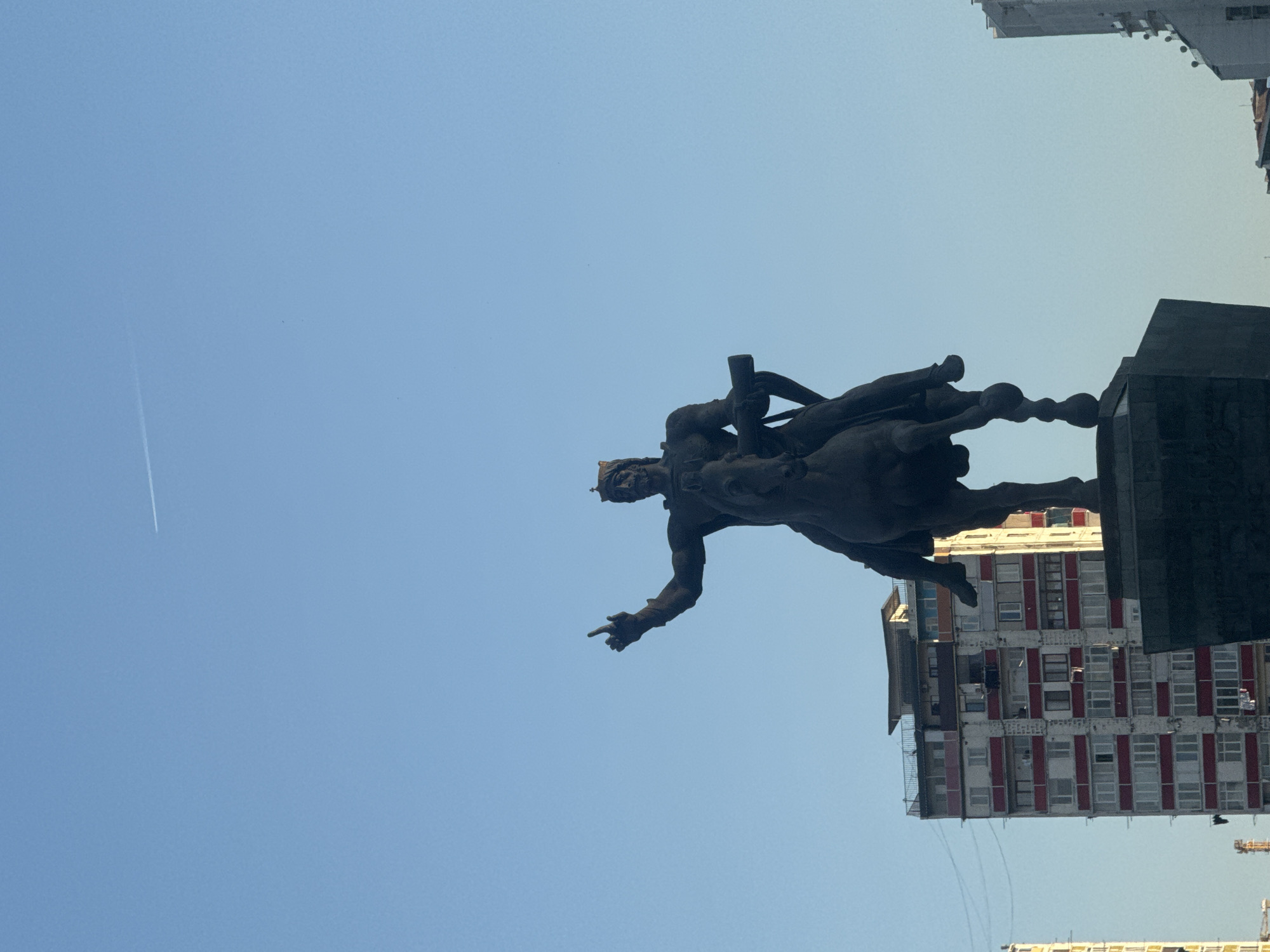 An upward-angled, daytime photograph features a prominent equestrian statue against a clear, bright blue sky in what is identified as Tbilisi, Georgia. The dark, monumental statue depicts a crowned male figure, likely King Vakhtang Gorgasali, mounted on a horse. The figure's right arm is outstretched with a pointing finger, while his left hand clutches a staff or weapon.
The statue is positioned towards the right side of the frame, partially silhouetted. In the background, to the right and above the statue, several modern buildings are visible. One notable building exhibits a facade with distinct red and white vertical stripes across its upper floors, suggesting balconies or structural divisions. Another building further back shows a more typical concrete and window design. A faint white contrail from an aircraft is subtly visible in the upper left section of the clear blue sky. The overall impression is a grand historical monument set against a contemporary urban backdrop under bright, fair weather conditions. No visible text is present in the image.