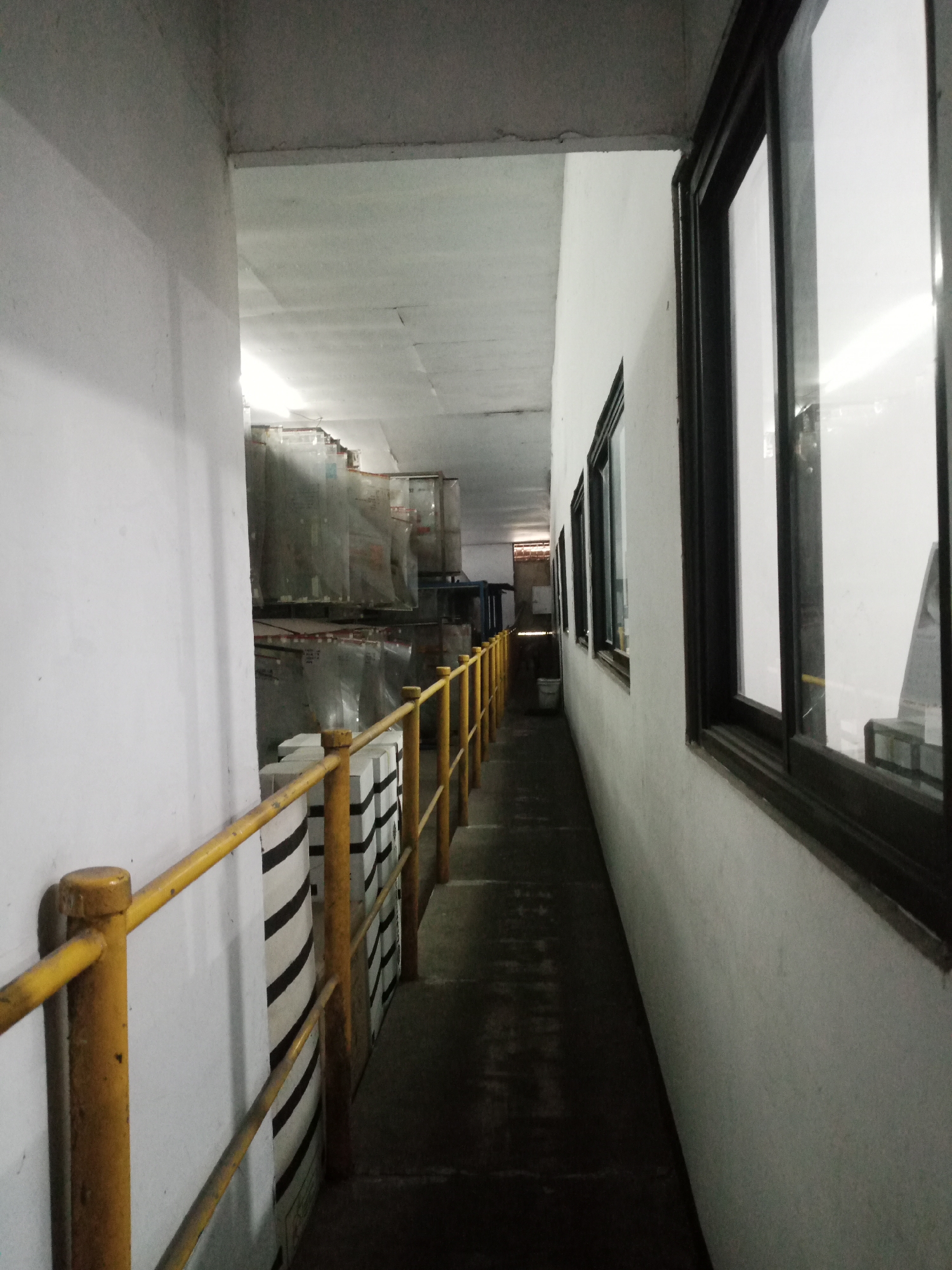 The image depicts a long, narrow hallway within an industrial or warehouse setting. The hallway is flanked by white walls on both sides. On the left, a yellow railing runs along the wall, which appears to be made of concrete. Beyond the railing, stacks of goods are visible, some covered in plastic sheeting. These items are arranged in rows and appear to be quite tall. On the right side of the hallway, a series of windows with dark frames offer glimpses into an adjacent space, which seems brightly lit. The floor of the hallway is dark and appears to be made of concrete or a similar material. The ceiling is high and made of corrugated material, with some visible lighting fixtures. The overall impression is one of a functional, possibly storage or transit area. The context of Balaraja, Indonesia, suggests this could be part of a factory, distribution center, or similar industrial facility located in that region. No people are visible in the image.