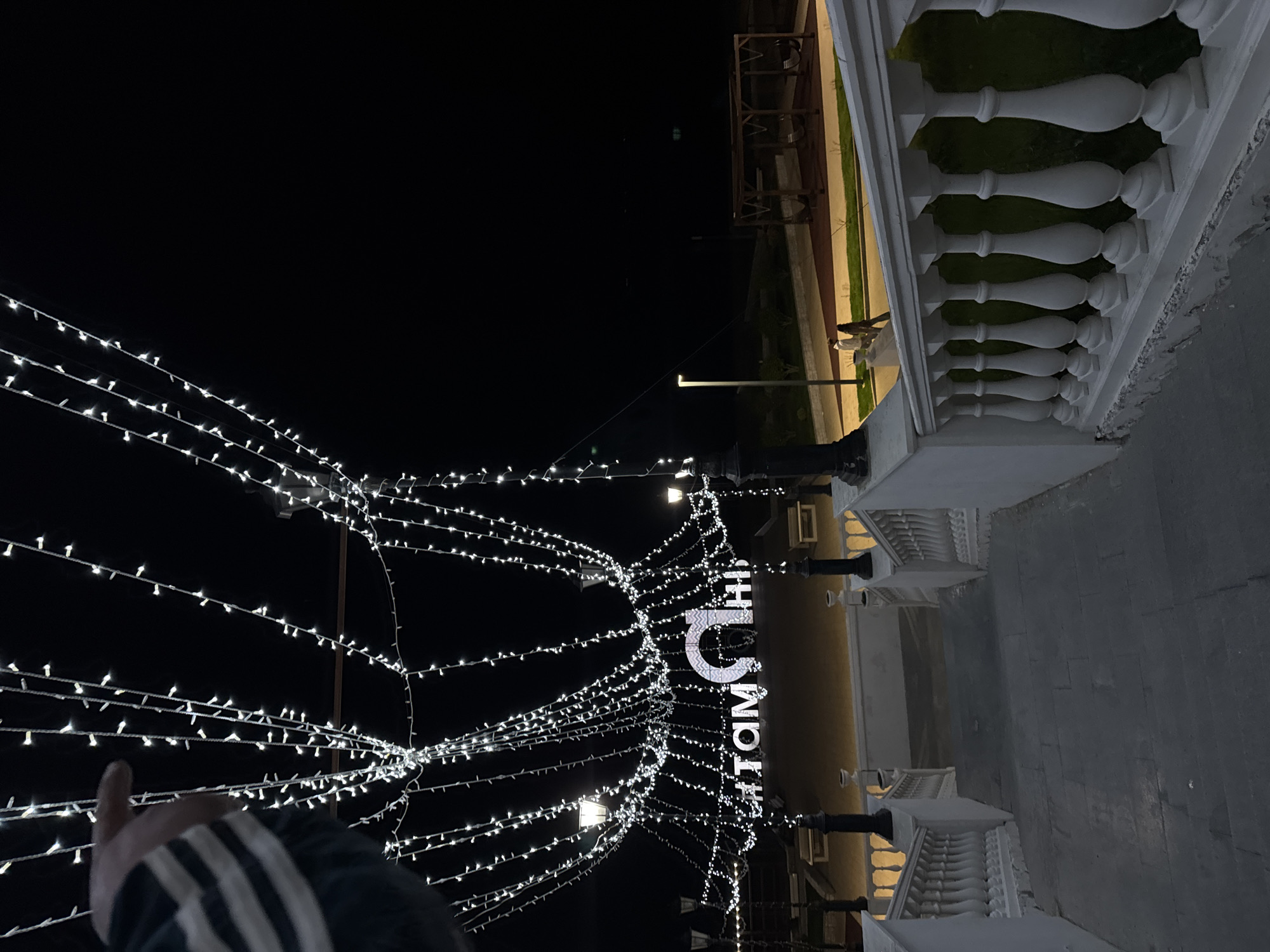 The image shows an outdoor scene in Taman, Russia, at night. The frame is filled with strings of white lights creating a festive atmosphere. The lights illuminate a structure with a sign written in Russian, which appears to read "ТАМАНЬ".
A white balustrade runs along the right side of the image, suggesting a balcony or elevated walkway. A person's hand and part of their striped sleeve are visible in the lower left corner. In the background, a few figures can be discerned walking on a pathway, and there are wooden structures that could be seating areas. The overall impression is that of an evening stroll in a decorated public space.