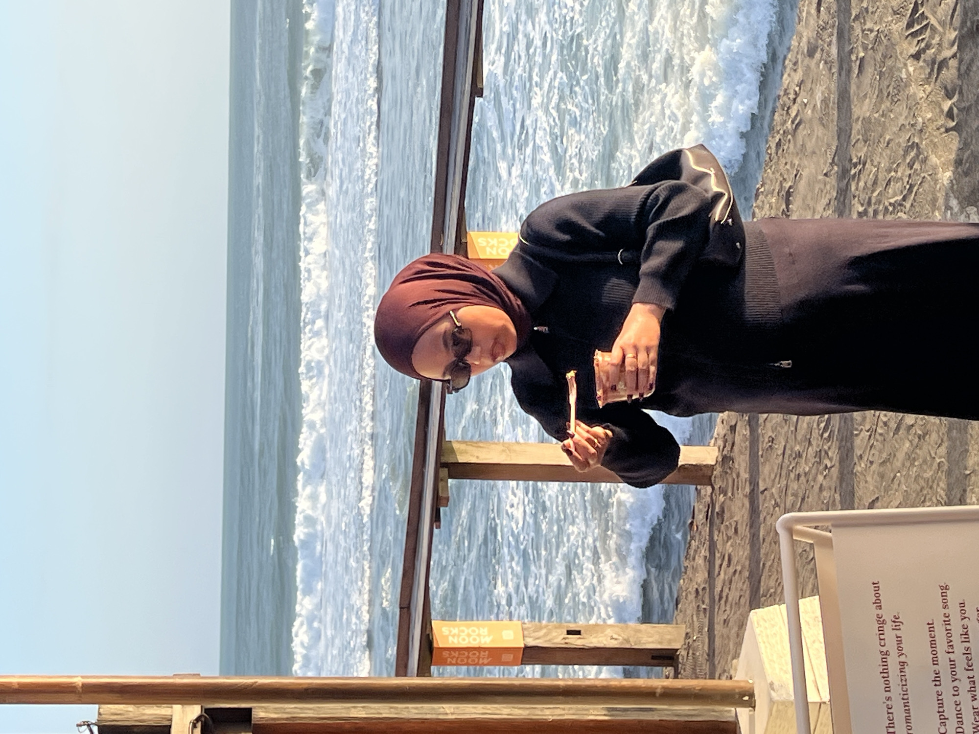A woman wearing a dark purple hijab and a dark long-sleeved dress, with sunglasses, stands on a sandy beach in North Kuta, Indonesia. She is holding a small cup and a spoon, appearing to eat from the cup. The ocean with waves is visible behind her, and a wooden structure with orange signage that reads "MOON ROCKS" is to her left. The lighting suggests it is late afternoon.