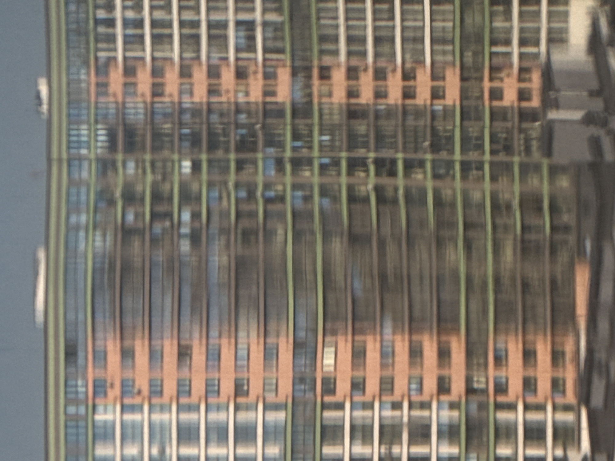 The image depicts a modern, multi-story residential building with a segmented facade. The building features a repeating pattern of windows and balconies. The left side of the building is clad in white and glass, while the central and right sections are characterized by terracotta-colored panels and darker glass. A prominent feature is the horizontal band of green that runs across multiple floors, separating different sections of the facade. The building appears to be partially in shadow, with sunlight highlighting certain areas and creating reflections on the glass surfaces. The overall impression is of a contemporary urban architectural style. The building is located in Minsk, Belarus, specifically at ul. Kurchatova, 1. The perspective suggests the photograph was taken from a distance, possibly from another building or across a street, giving a broad view of the building's exterior. No people are visible in the image.