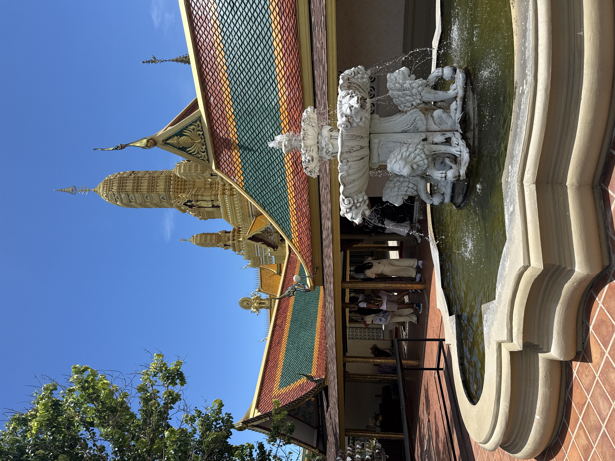 The image shows a low-angle view of Wat Asokaram in Bang Pu Subdistrict Municipality, Thailand. A multi-tiered golden pagoda with elaborate spire rises against a clear blue sky, partially obscured by the tiled roof of a nearby building. The roof is adorned with colorful tiles (green, orange, and red) and intricate gold trim. A multi-tiered white stone fountain with lion head spouts is situated beside the building. Water is actively flowing from the fountain into a stone basin containing dark water. Behind the building’s entrance, several people are present, some appearing to wear light-colored clothing and stand near a railing. A tree with green foliage is visible in the lower left corner of the frame. The foreground shows the tiled exterior of the building, providing a close perspective of the temple's architectural elements.
