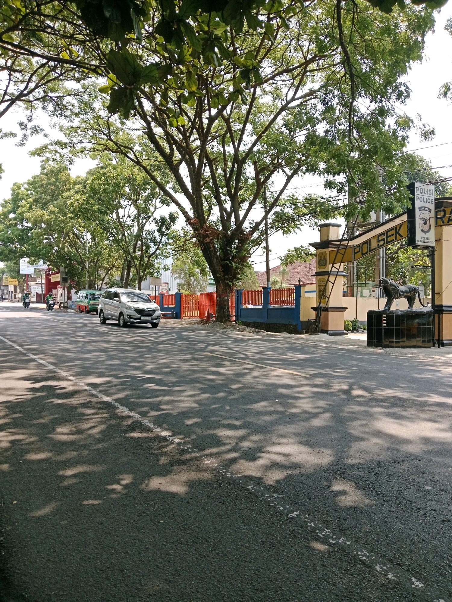 In Karangtengah, Indonesia, a street scene unfolds beneath the dappled shade of mature trees. The asphalt road, marked with a white dashed line, runs towards the right of the frame. On the right side of the road, a police station, identified by a sign reading "POLSEK" and further text in Indonesian, is prominent. The police station features an arched entrance with ornate pillars and a black lion statue at its base. A blue and red fence borders the police station. Across the street, a silver car drives away from the camera, and two motorcycles, one carrying a rider and the other a passenger, are also present on the road. A colorful miniature bus, likely a public transport vehicle, is parked further down the street on the left. The environment suggests a sunny day with a light breeze, as evidenced by the moving leaves and the shadows cast on the road.