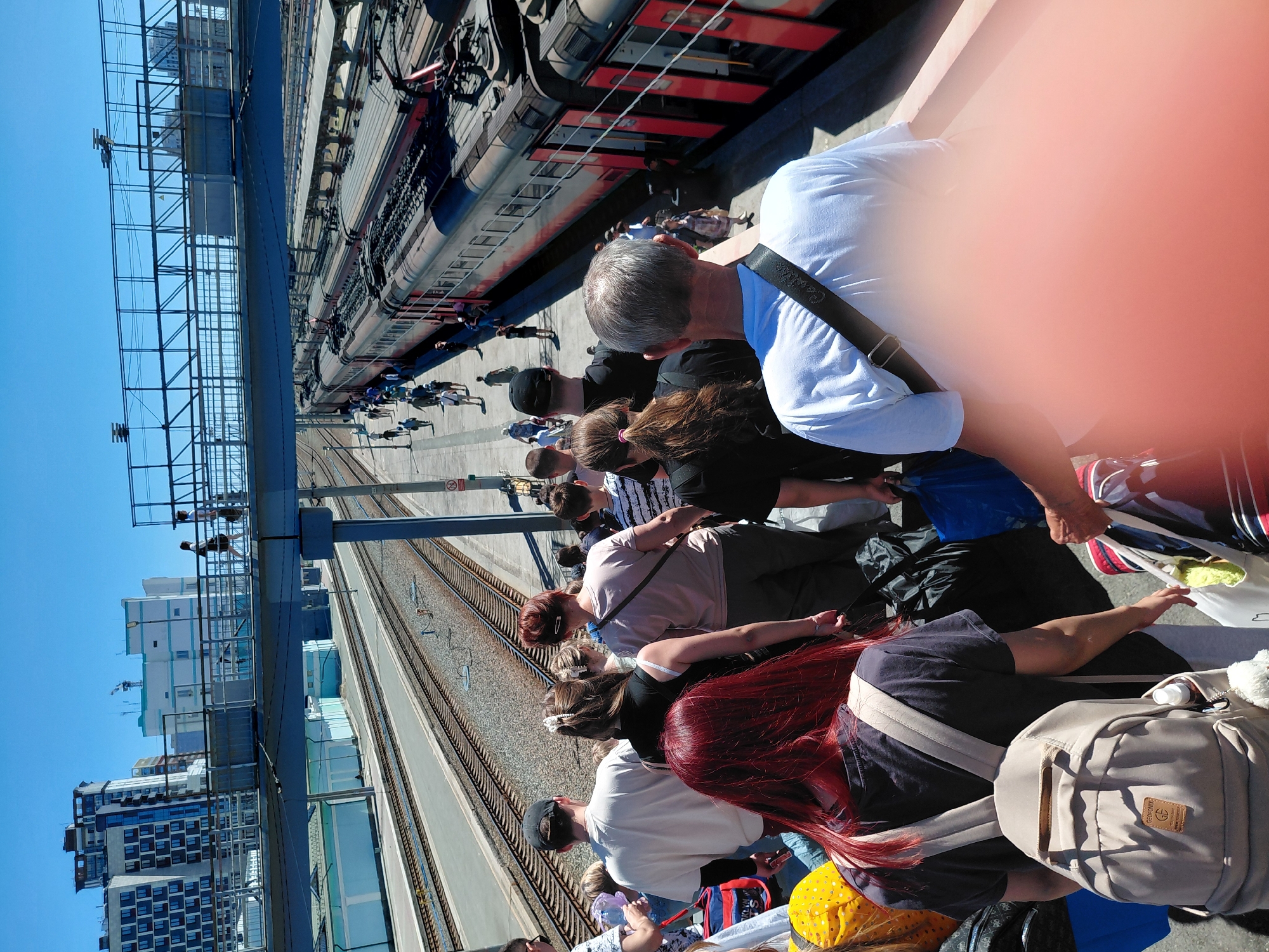 In the city of Unknown, Unknown, on a sunny day, a group of people are seen on a pedestrian bridge above a train station. The bridge overlooks several train tracks and passenger trains. Many individuals are walking in the same direction, some carrying luggage, suggesting they are either arriving at or departing from the station. The environment appears to be an urban train station with modern buildings in the background. The scene captures a moment of transit for the travelers.