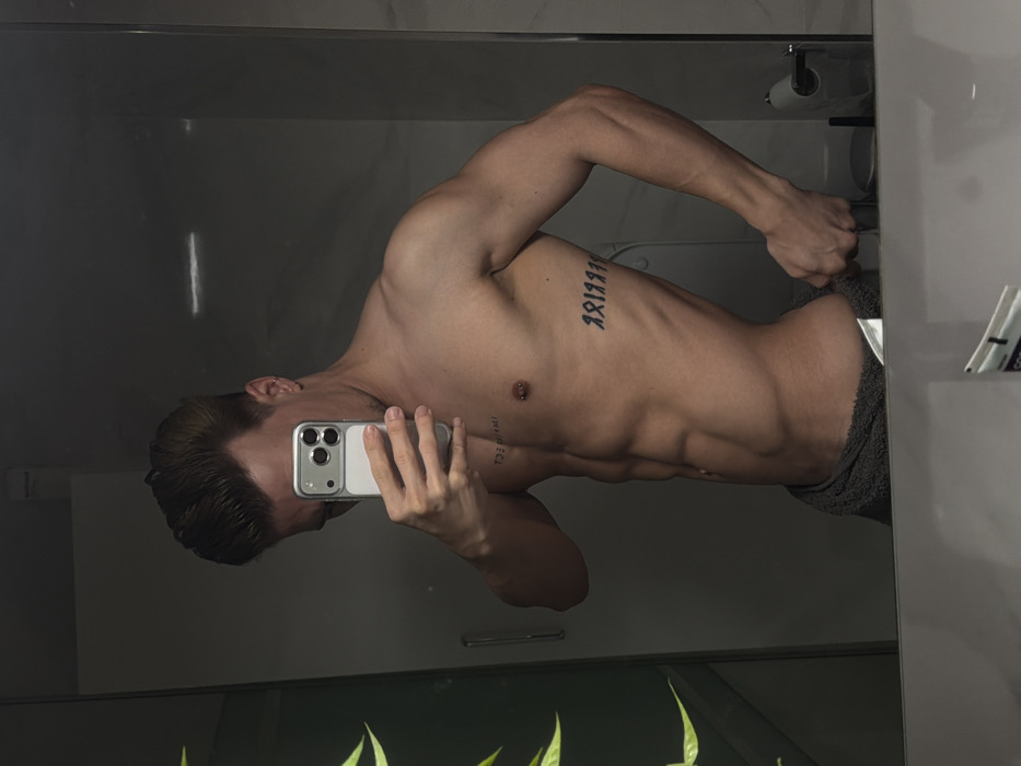 An adult male is captured in a mirror selfie within a modern bathroom, presumed to be located in Viseu, Portugal. The subject, who has a lean, muscular physique and short brown hair styled upwards, is bare-chested. He holds a dark grey towel around his lower torso with his right hand, which is visible gripping the towel's edge. He is taking the photograph with a silver-cased smartphone, equipped with a triple camera array, held by his left hand, which partially obscures his face. Distinctive body markings include a vertical chest tattoo on his left pectoral muscle, appearing to read "IMPERFECT" (reversed due to reflection), a horizontal tattoo of symbolic characters on his right rib cage, and a small barbell piercing on his right nipple. The environment, reflected in the mirror, features white tiled walls, a door handle or vertical fixture on the left, a toilet paper roll dispenser, and part of a toilet on the far right. A soft green reflection, possibly from foliage or a plant, is visible on the left side of the mirror.