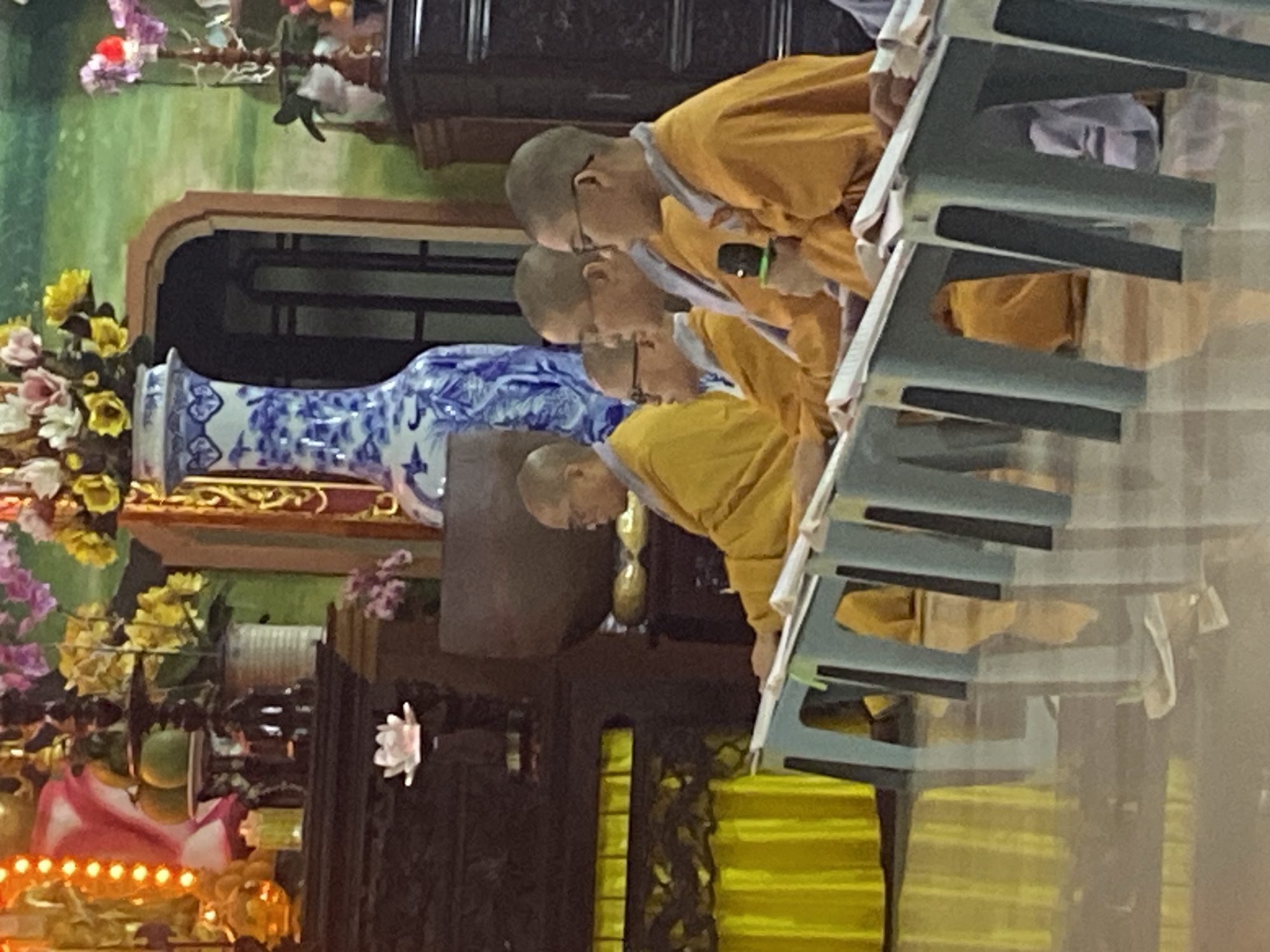 A group of Buddhist monks in saffron robes are seated in a row at low tables, facing right, in what appears to be a temple or meditation hall in Phu Thuy Ward, Vietnam. The monks are all wearing glasses and have shaved heads. The monk in the foreground is holding a microphone. The tables have books or scriptures on them. In the background, there are decorative floral arrangements and a large blue and white vase. The floor is reflective, suggesting it may be polished tile or marble.
