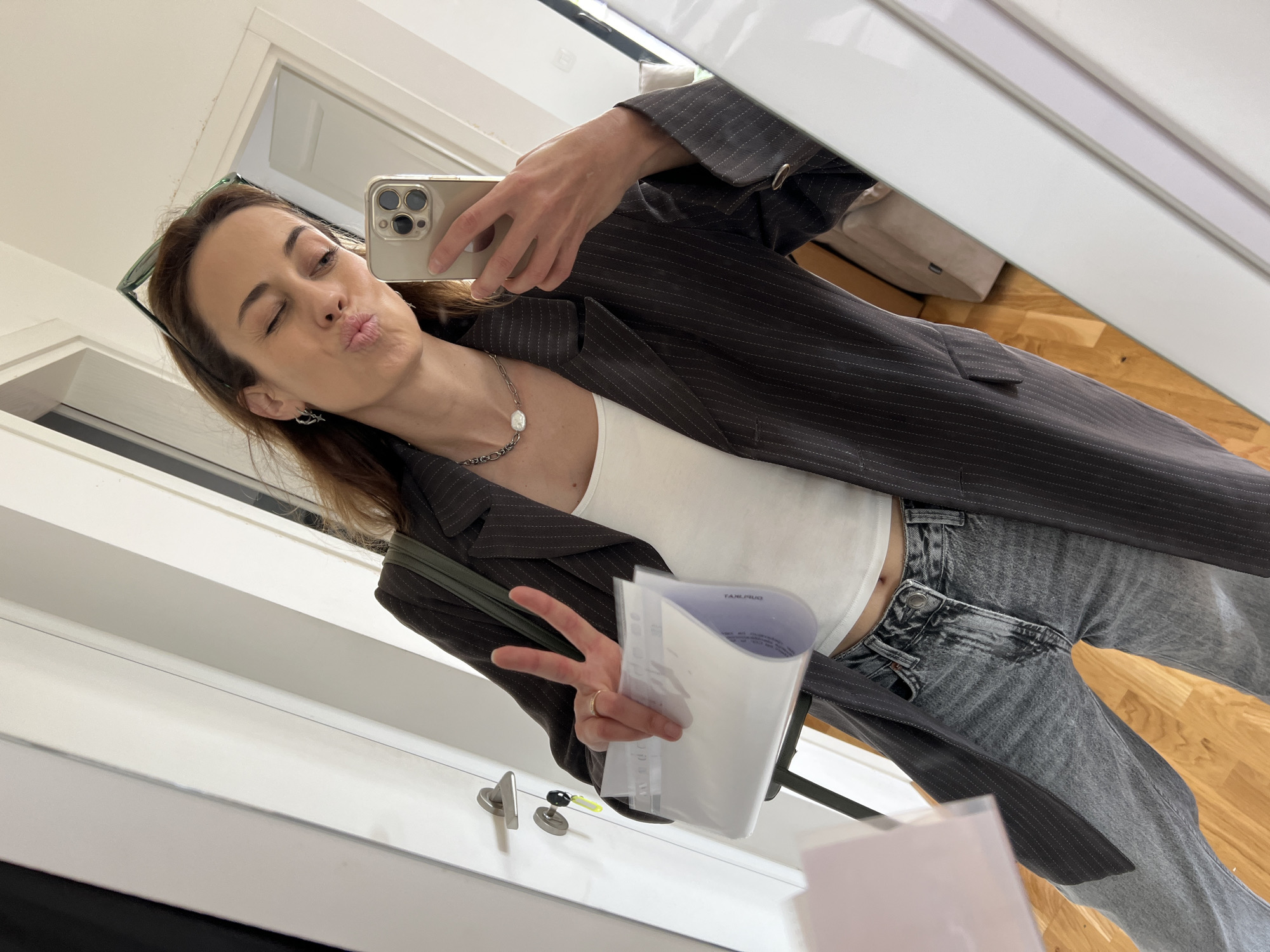 A woman is depicted taking a mirror selfie indoors, in what appears to be a hallway or room in Bar, Montenegro. She is dressed in a dark pinstriped blazer over a white crop top, paired with light-wash, high-waisted jeans. Sunglasses are perched on her head, and she wears a necklace with a pearl pendant and an earring.
Her left hand holds a light-colored smartphone, capturing her reflection. With her right hand, she holds a clear plastic document holder containing papers and simultaneously makes a peace sign. She is looking directly at the phone's camera, with a playful expression: puckered lips in a kissing gesture and one eye winking.
The setting consists of white walls and door frames, with a wooden floor visible in the reflection. Door handles are noticeable on a white door in the lower part of the frame. Bright lighting suggests it is daytime. On the papers inside the clear document holder, partially visible, reversed text reads "TAXLЯUG" (likely "TAXIRUG" or similar). The overall mood conveyed is casual and cheerful.