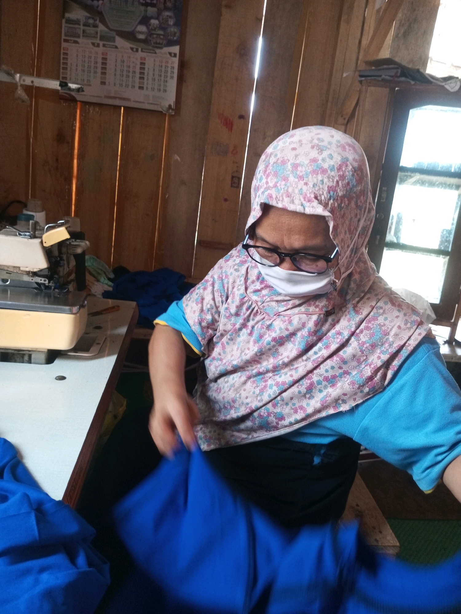 In Kalibeber, Indonesia, a woman is seen from the chest up, wearing a floral-patterned headscarf and a face mask. She is also wearing glasses and a blue long-sleeved shirt with a yellow trim. Her hands are in motion, holding a piece of blue fabric. She appears to be working at a sewing machine, which is visible on a white table to her left. The background consists of rustic wooden walls, suggesting a workshop or a similar domestic setting. A calendar hangs on the wall behind her. The lighting is dim, with sunlight filtering in from a window on the right side of the frame.