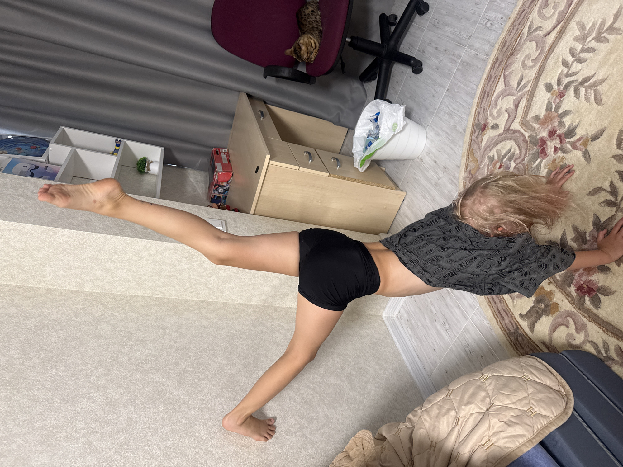 A young girl with blonde hair is captured in a dynamic pose on what appears to be a textured carpet. She is wearing a dark, short-sleeved, open-weave top and black shorts. Her left leg is extended straight out behind her, parallel to the floor, while her right leg is bent at the knee, supporting her body. Her arms are reaching out to her sides, with her right hand resting on the carpet.
The background reveals a domestic setting. To the left, there are white shelves containing books or toys, and a grey pleated curtain. A desk chair with a maroon seat and a black base is visible, with a cat lounging on the seat. A light-colored wooden cabinet with drawers stands next to the chair, and a white trash can with a plastic bag is placed nearby. The floor is tiled and there is a large, ornate rug with floral patterns. To the right, a quilted beige blanket covers part of a dark blue object, possibly a piece of luggage.
The lighting suggests it is daytime, but the exact time and weather are not discernible. The girl's pose suggests she is engaged in physical activity, possibly gymnastics or dance. Her expression is not visible. There is no visible text in the image.