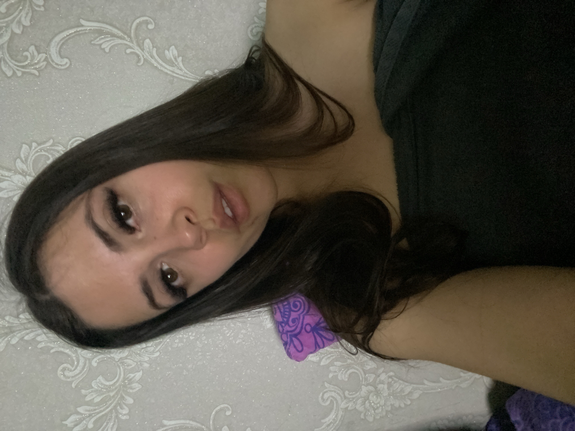 A young woman with long, dark hair is lying on a bed with a patterned white and silver headboard, presumably indoors. She has fair skin, dark eyebrows, and full lips. She is wearing dark clothing. Part of a purple pillow is visible near her head. The image's location within the city of Chaikovsky, Russia, cannot be determined from the available visual information.
