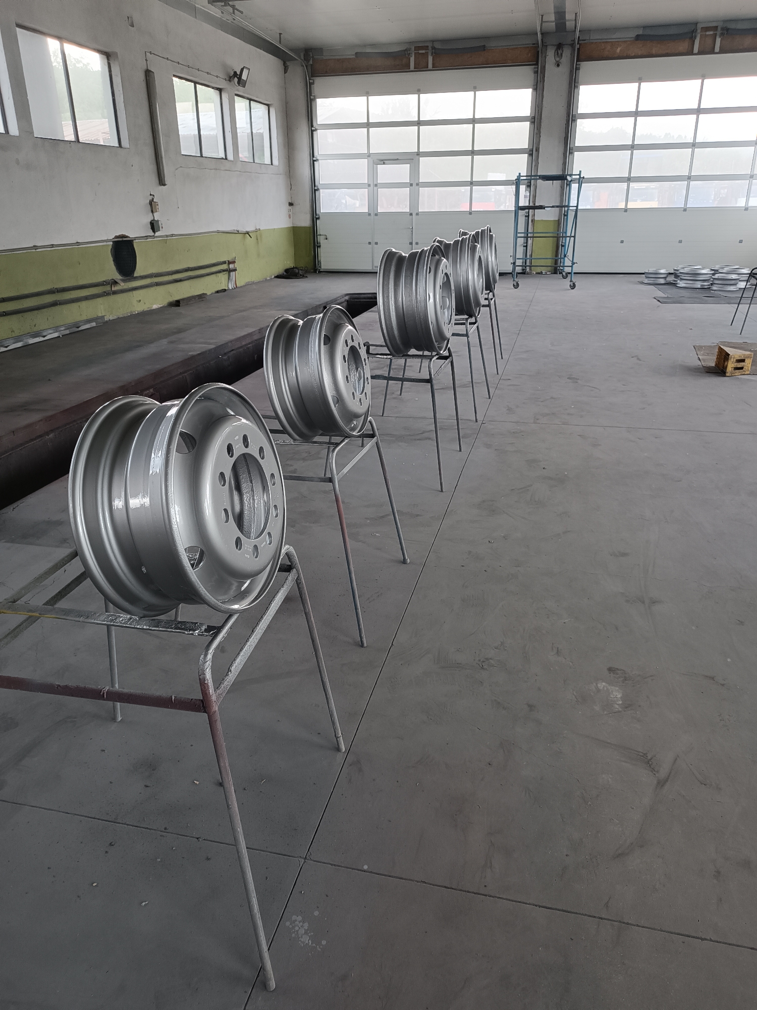 This media file presents an interior view of a large industrial workshop or garage located in Dobrodan, Bulgaria. The primary subjects are eight large, silver-grey metal wheels, likely for trucks or heavy vehicles, arranged in a line towards the center and right of the frame. Each wheel, appearing freshly painted or refurbished with a uniform matte metallic finish, rests on its own simple, slender metal stand, suggesting a stage in a drying, inspection, or processing operation.
The setting is an expansive, high-ceilinged industrial space. On the left, a concrete wall is painted white on top with a faded olive-green band along the lower section. Several large, rectangular windows punctuate this wall, allowing diffused natural light to illuminate the interior. Below the windows, a recessed area or pit, possibly for vehicle maintenance, is visible. On the right and rear of the image, two large, white sectional garage doors dominate, providing additional ample natural light and a soft, hazy view of the outside environment, hinting at trees or distant structures.
In the mid-ground, to the right of the row of wheels, a blue metal scaffolding unit on wheels is visible. Further into the background, stacks of round metal objects, possibly more wheels or components, are laid out on the floor, alongside a wooden pallet. The floor throughout the workshop is made of grey concrete, showing typical signs of wear, scuffs, and expansion joints.
The bright, yet soft and diffused, natural light streaming in from the windows and garage doors indicates that it is daytime. There are no people visible in the image, and no discernible text can be read on any objects or surfaces. The overall impression is one of an active, functional industrial environment focused on vehicle component maintenance or manufacturing.