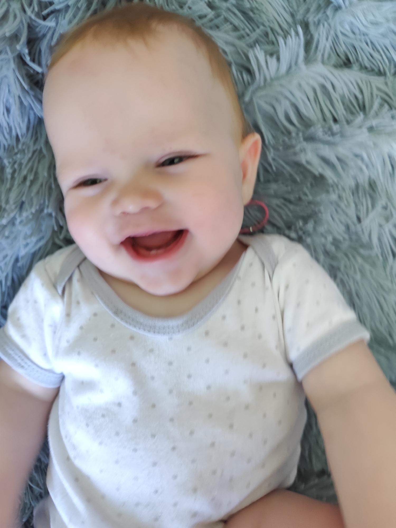 A close-up shot features a baby lying on a fluffy, bluish-gray blanket. The baby has fair skin, reddish-blonde hair, and a wide, toothless smile, revealing their open mouth. They are wearing a white onesie with light gray trim and small gray dots. A red hair tie is visible near the baby's right ear. The baby's limbs extend into the foreground. The location in the city of Фоки, Russia, cannot be determined from the image.