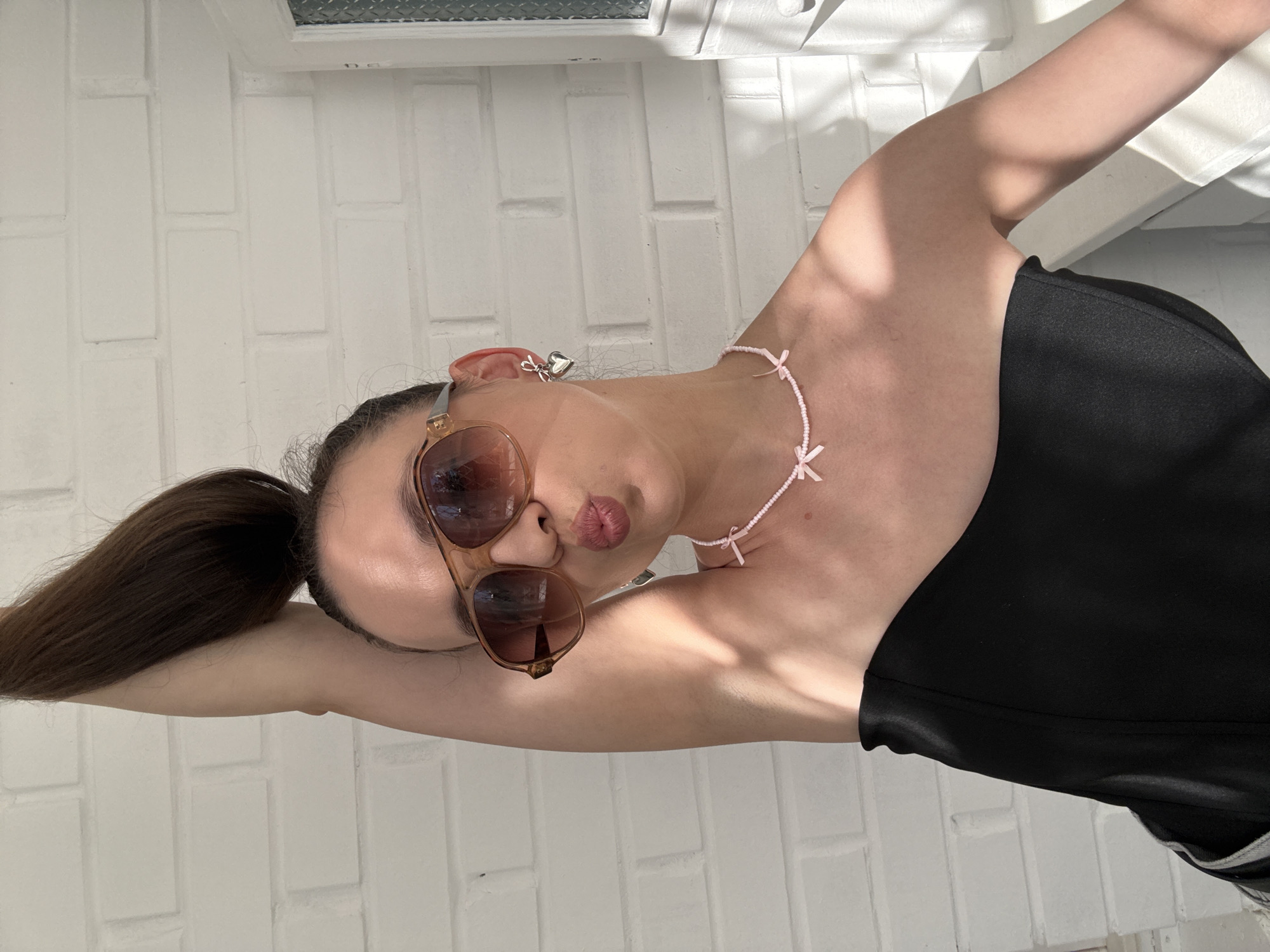 A person is lying down, with their head resting on their arm, in front of a white brick wall. They are wearing sunglasses and a black top. A delicate pink beaded necklace with tiny bows is visible around their neck. The person has dark hair pulled into a ponytail. The lighting suggests it is daytime, with shadows cast on the wall, indicating sunlight. The overall mood is relaxed and casual. There is no visible text in the image.