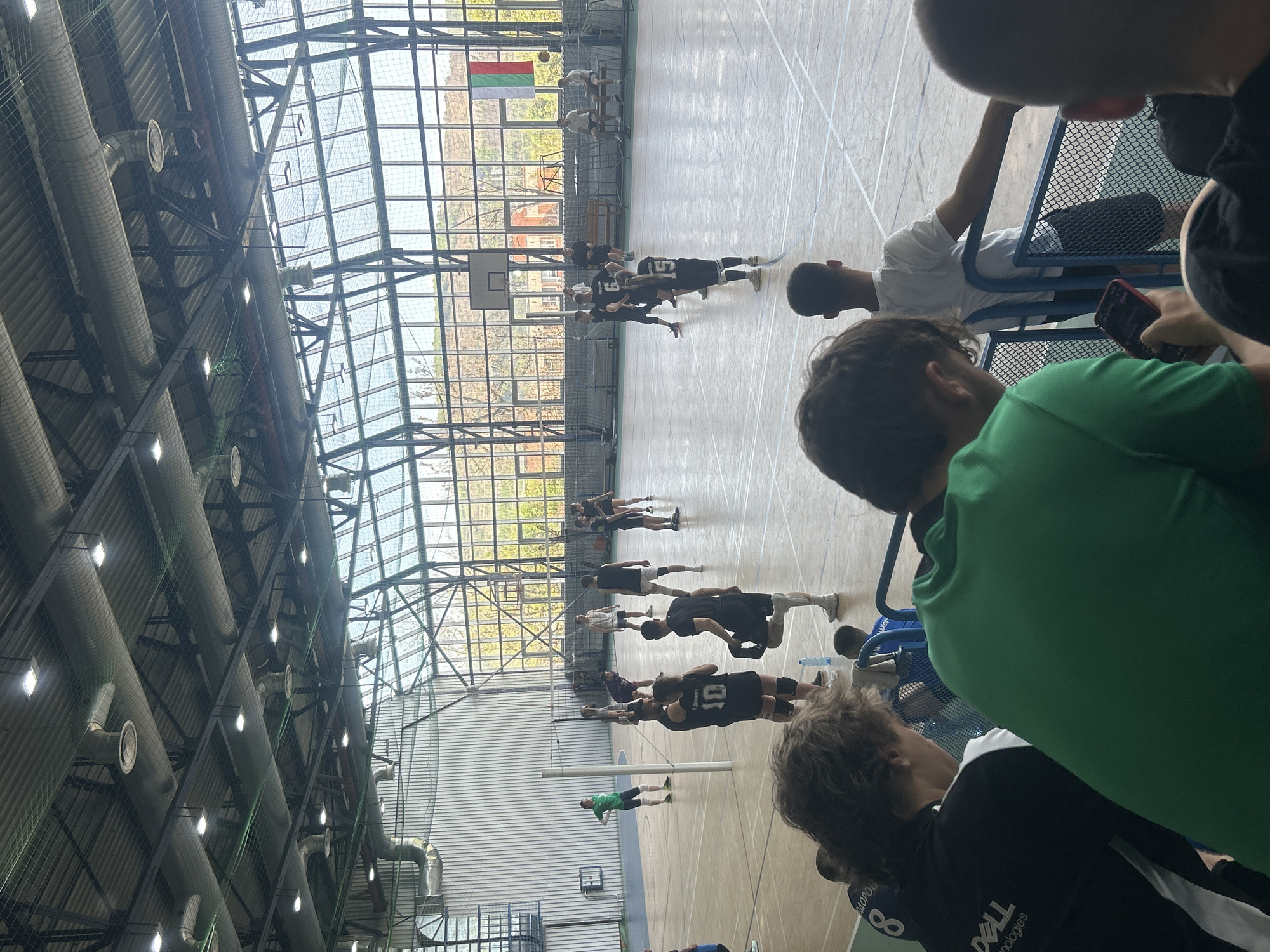 The image depicts an indoor sports hall, likely in Veliko Tarnovo, Bulgaria, as evidenced by the prominently displayed Bulgarian flag (white, green, red horizontal stripes) hanging high on a wall.
In the foreground and to the right, several individuals are observed from behind or the side, seated on blue metal chairs, acting as spectators or waiting participants. One person is seen looking at a red cell phone. Visible clothing includes a green athletic shirt and a black and white shirt with "DELL Technologies" printed on the back.
The central activity takes place on a light-colored wooden court, marked with lines for sports. A group of young men, dressed in athletic wear, primarily black jerseys with white numbers, are engaged in what appears to be a volleyball practice or game. One player's jersey clearly shows "TSVETKOV" above the number 10. A volleyball net is visible, along with a basketball hoop in the background, indicating a multi-purpose gymnasium. A single player in a green shirt stands further down the court.
The background features large, expansive windows that allow natural daylight to flood the hall, offering a glimpse of outdoor trees and distant buildings. The ceiling structure is industrial in design, with intricate metal framework, large ventilation ducts, and numerous bright, rectangular light fixtures. A protective netting is stretched across parts of the ceiling. The scene portrays an active daytime sports environment.
