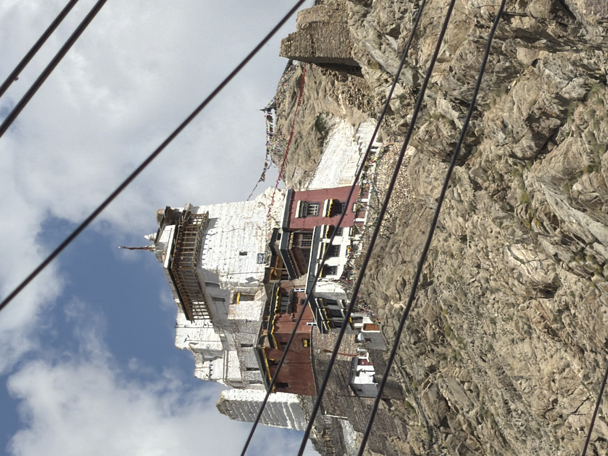 The image shows the Tsemo Monastery complex in Leh, India, perched on a rocky hillside. The monastery is a multi-level structure with white-washed buildings, featuring red-painted sections and wooden accents. Prayer flags are strung across the hillside and between buildings. The foreground is dominated by a rocky, barren slope, with several black power lines running diagonally across the frame, obscuring parts of the view. The sky above is partly cloudy, with a mix of blue sky and white clouds. There are no people visible in the image. The overall scene depicts a remote, mountainous religious site under a bright sky.
