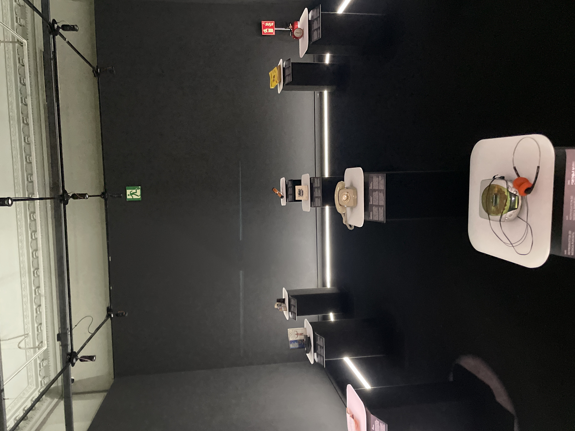 The image depicts an exhibition hall with a dark, minimalist aesthetic. The floor is highly reflective, creating a mirror-like surface. Several display pedestals of varying heights are arranged throughout the space, each topped with white rectangular platforms showcasing various objects.
On the left, a low, illuminated white display platform is partially visible. Further back, two dark pedestals stand side-by-side. The left pedestal has a white top with a book and other small objects. The right pedestal has a similar white top with two stacked items.
In the center of the exhibition, a dark pedestal supports a white platform with a vintage cream-colored telephone. To the right of this, another dark pedestal holds a white platform with what appears to be a yellow document. Next to it, a slightly taller dark pedestal displays a red alarm clock with a fire extinguisher symbol above it.
Further to the right, another display area is partially visible. In the foreground of the image, a white platform displays a portable music player with headphones. The walls are dark gray, and subtle uplighting emanates from the floor, highlighting the edges of the display platforms. The ceiling is white with intricate decorative molding and a track lighting system with several spotlights. A green emergency exit sign with a running figure is visible on the upper wall.
While the image is labeled as being in Barcelona, Spain, there are no specific visual cues within the photograph to confirm this location.